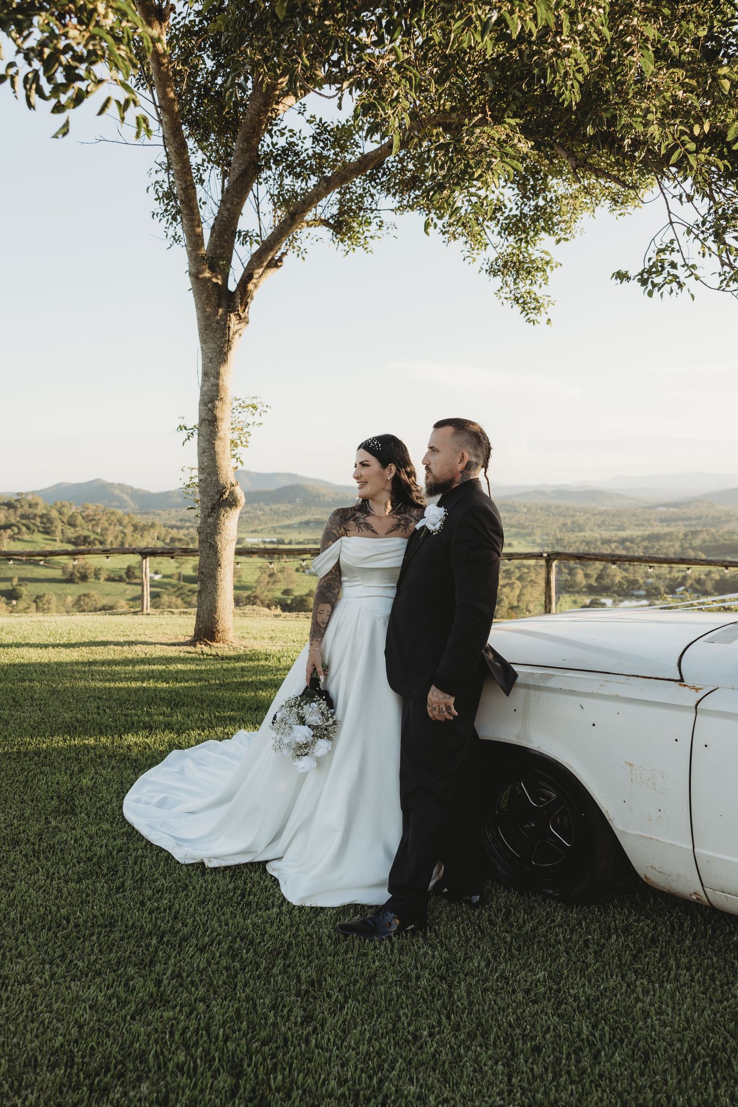 Newlyweds pose for romantic photos next to a vintage white car on a grassy hillside.