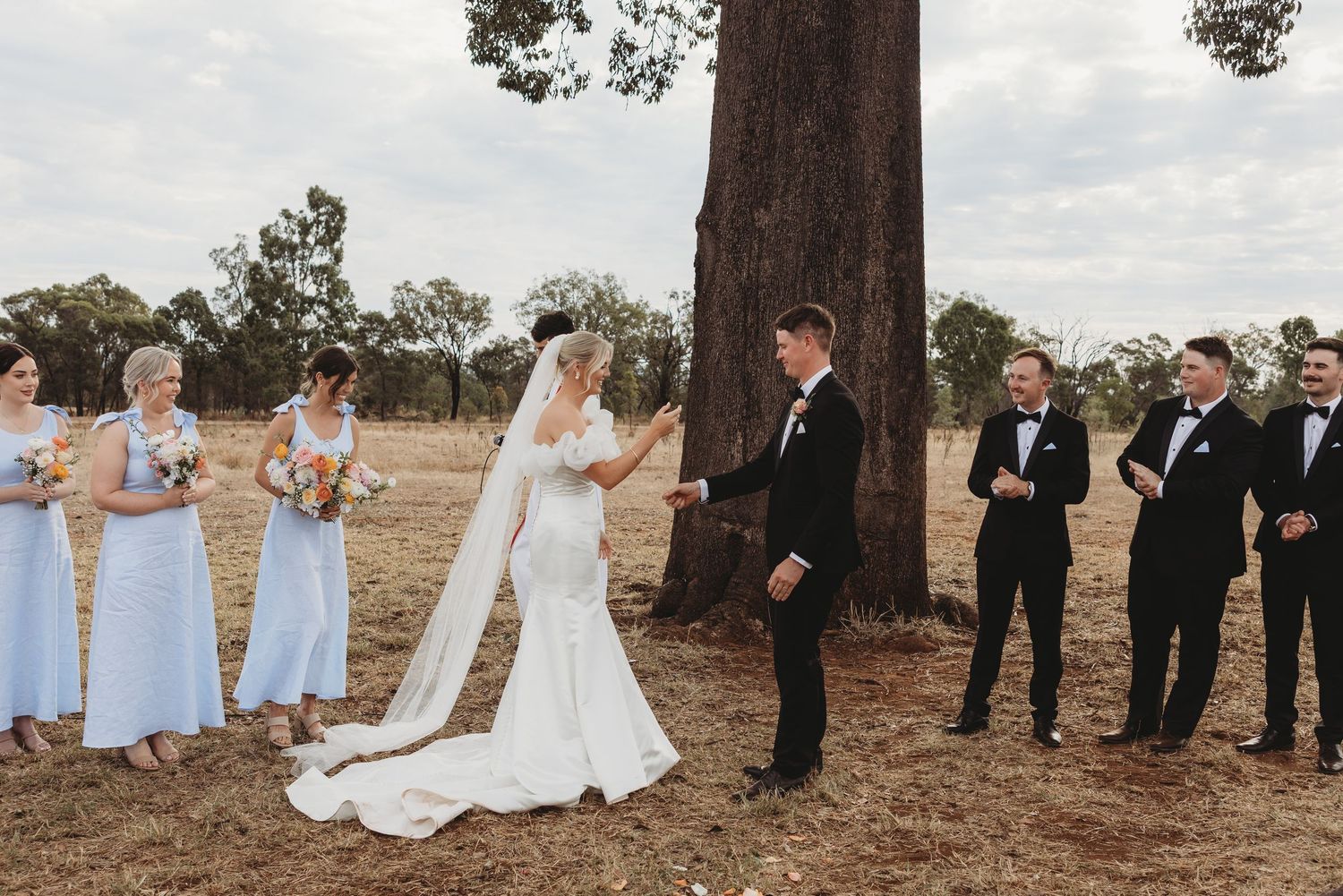 Wedding party lined up under large tree for formal group photos in countryside setting.