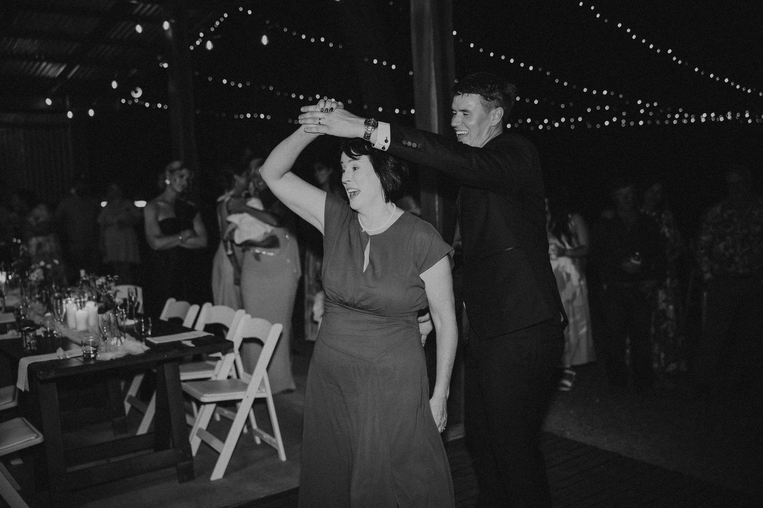 Wedding guests dancing at reception under string lights in black and white photography.