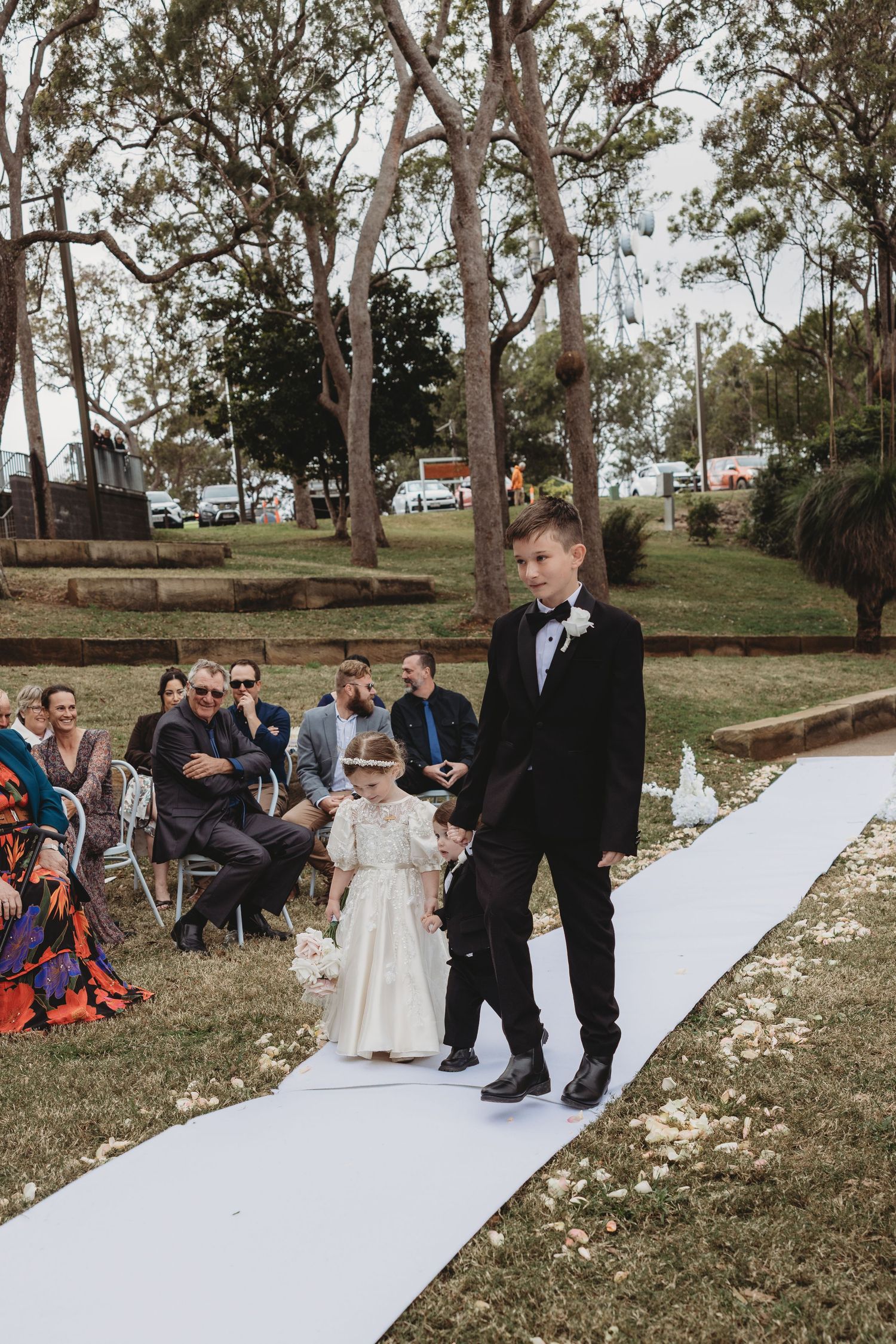 Wedding ceremony guests watch as couple walks down white aisle runner in outdoor garden setting.