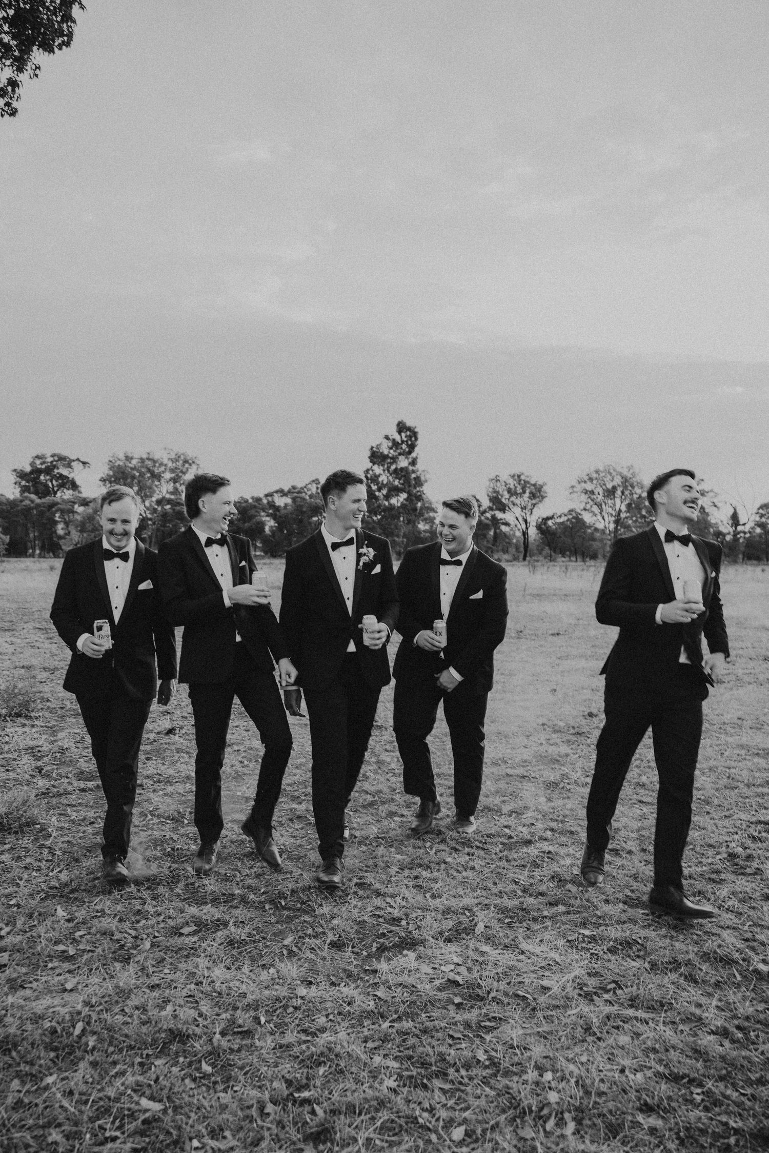 Groomsmen in black suits walk in formation across a grassy field in black and white photography.