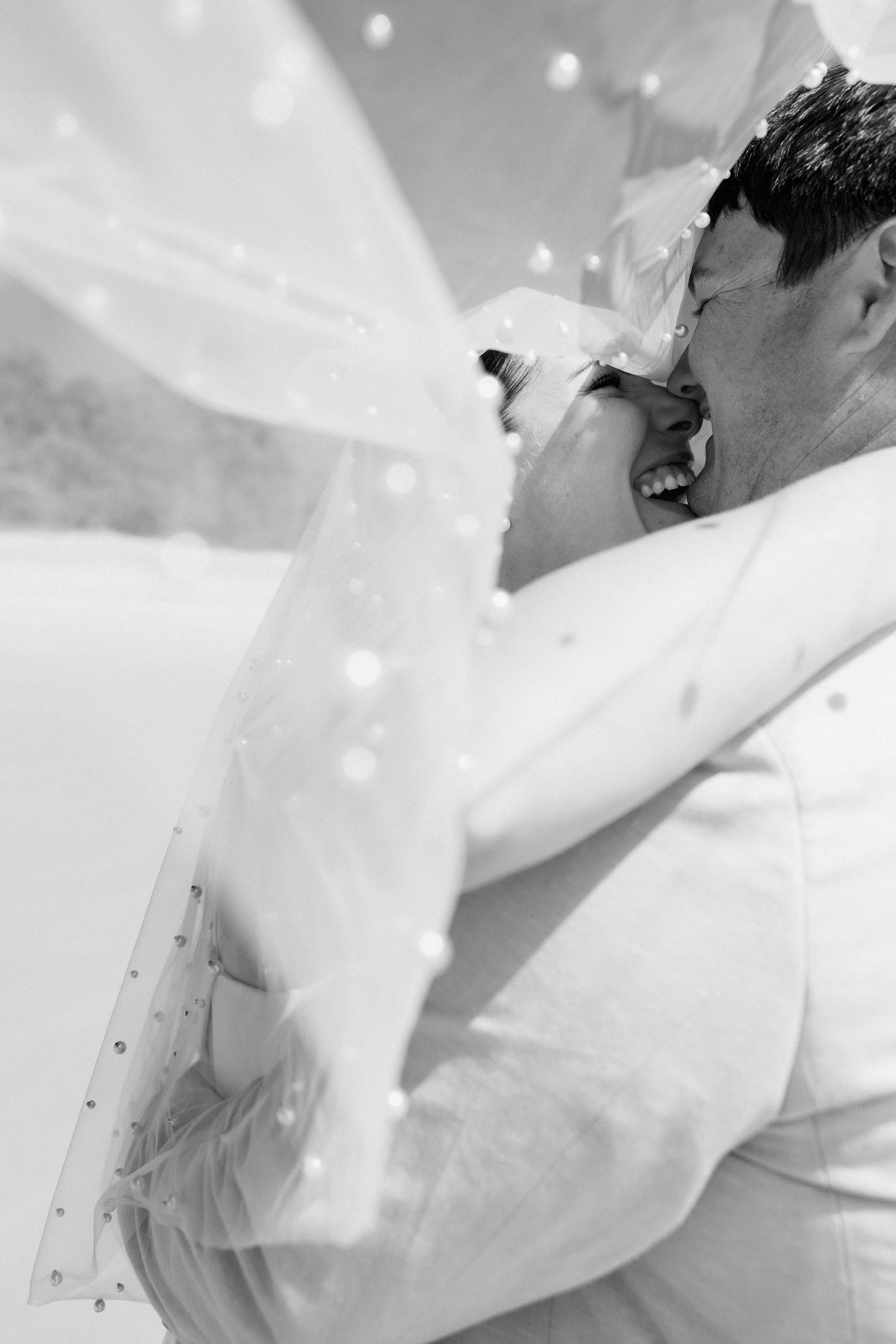 Black and white photo of a couple embracing on a beach, bride's veil blowing in the wind.