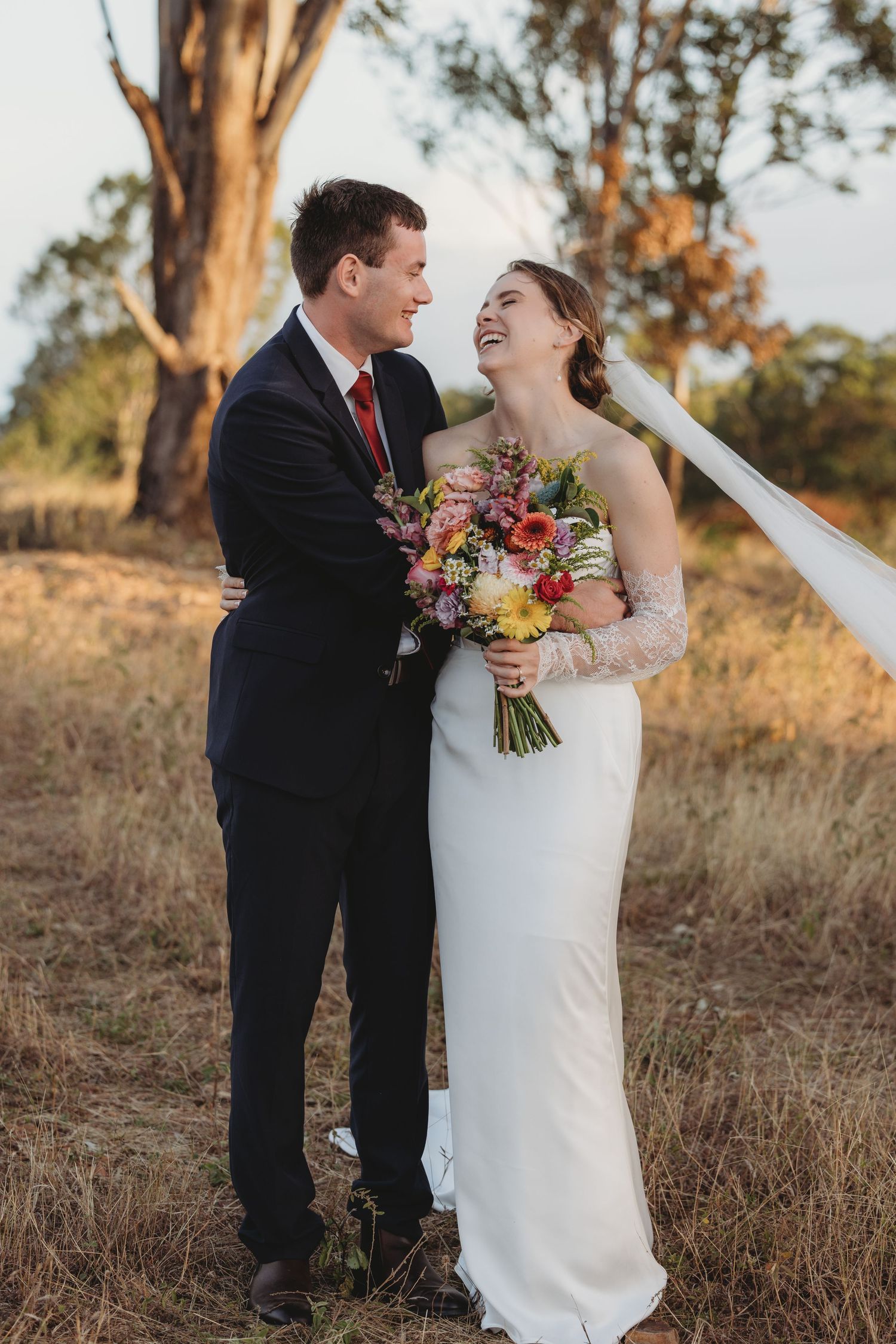 Laughing bride and groom pose in golden field holding colorful bouquet at sunset wedding.