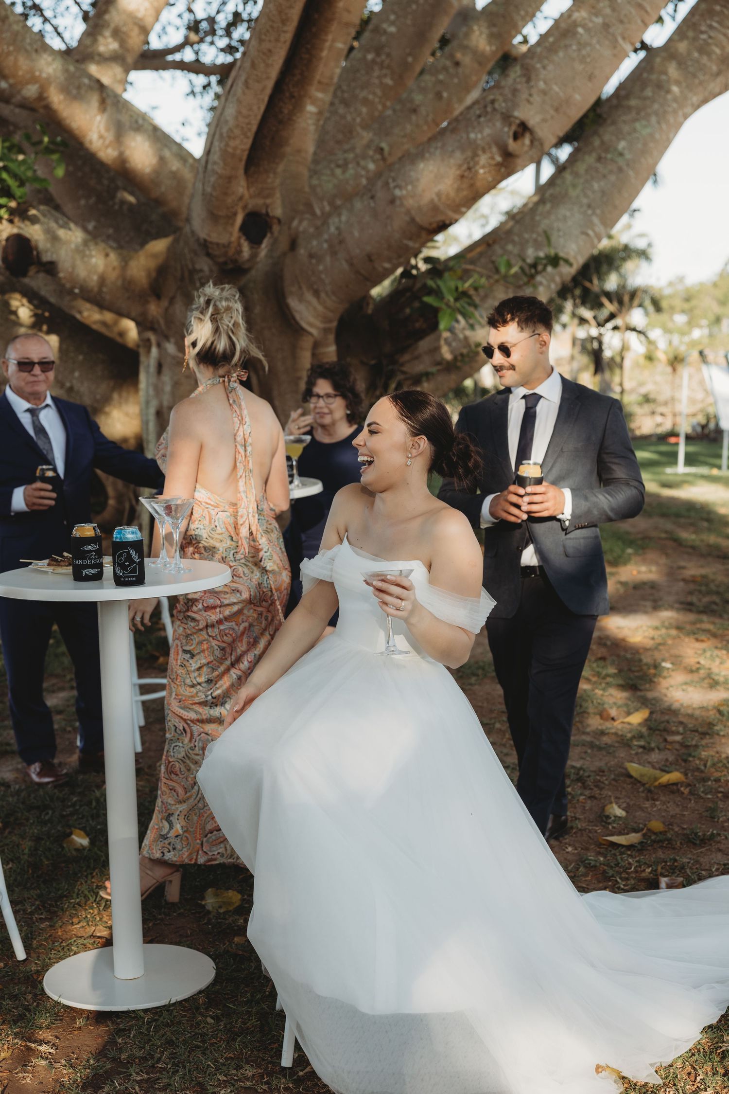 Wedding guests gather under large trees during an outdoor cocktail reception.