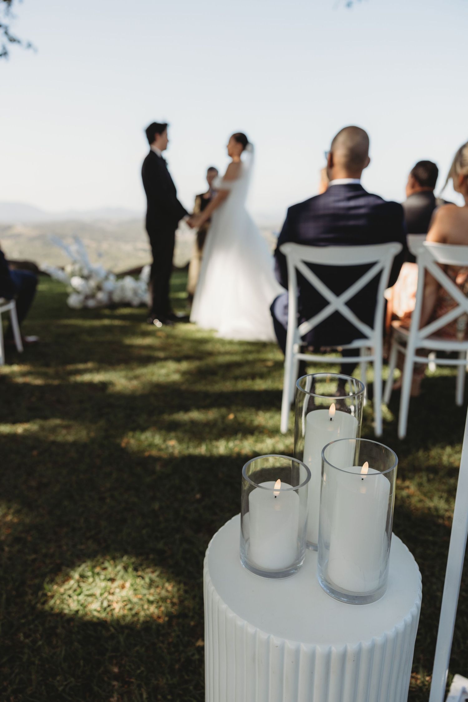 White candles in glass holders set up along the wedding ceremony aisle with guests in background.