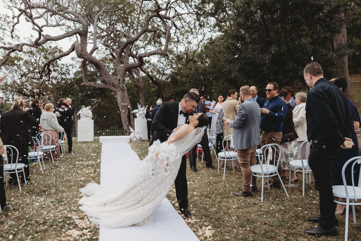 A couple shares a romantic dip and kiss during their outdoor wedding ceremony surrounded by guests and rustic chairs.