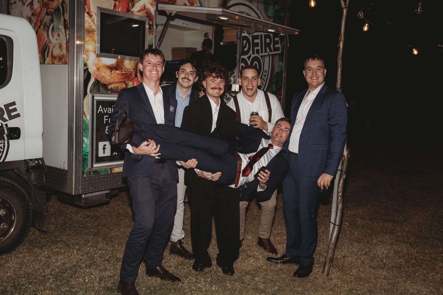 Groomsmen playfully lift groom outside food truck at fun outdoor wedding reception.