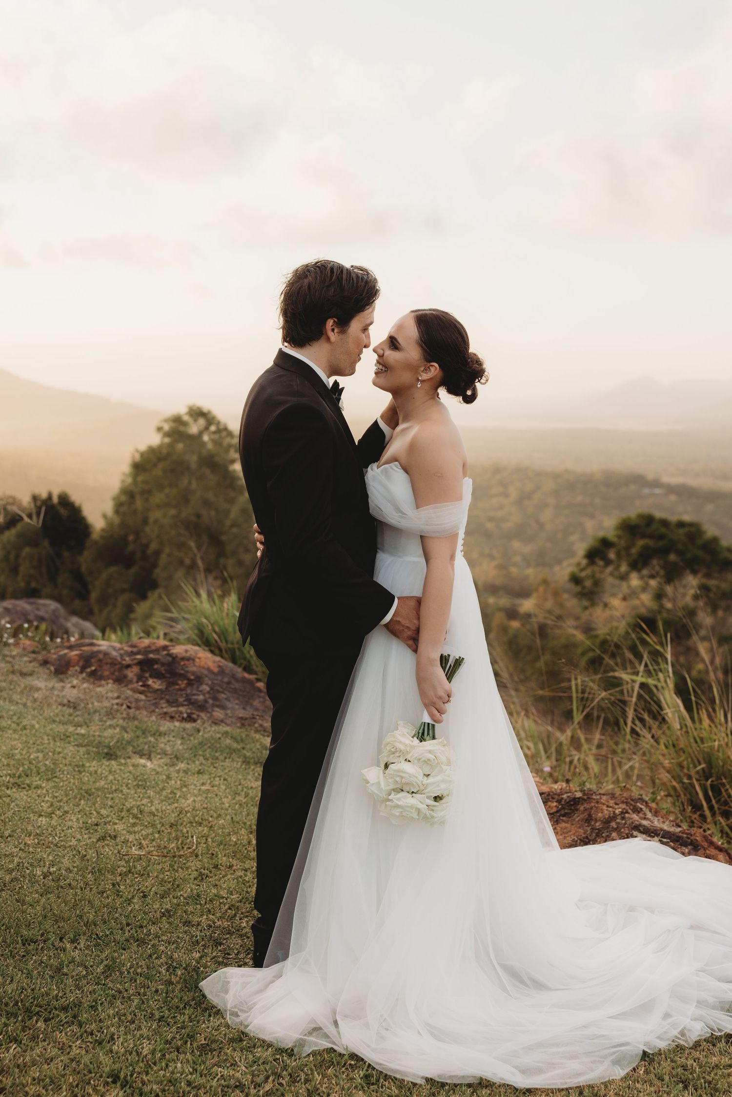 Bride in flowing white gown holds bouquet while kissing on scenic overlook.