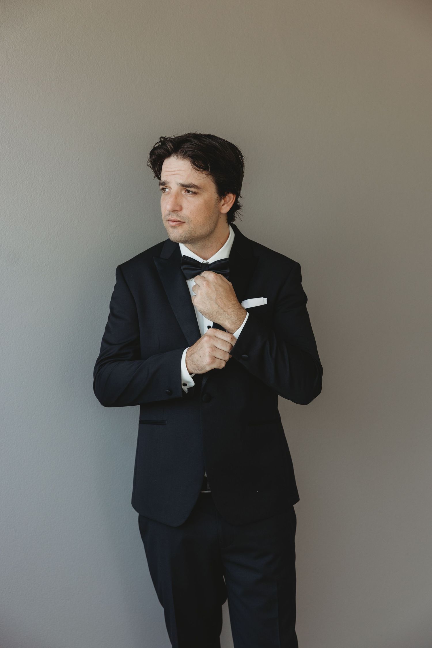 A person in a black tuxedo poses against a grey wall in various contemplative stances.