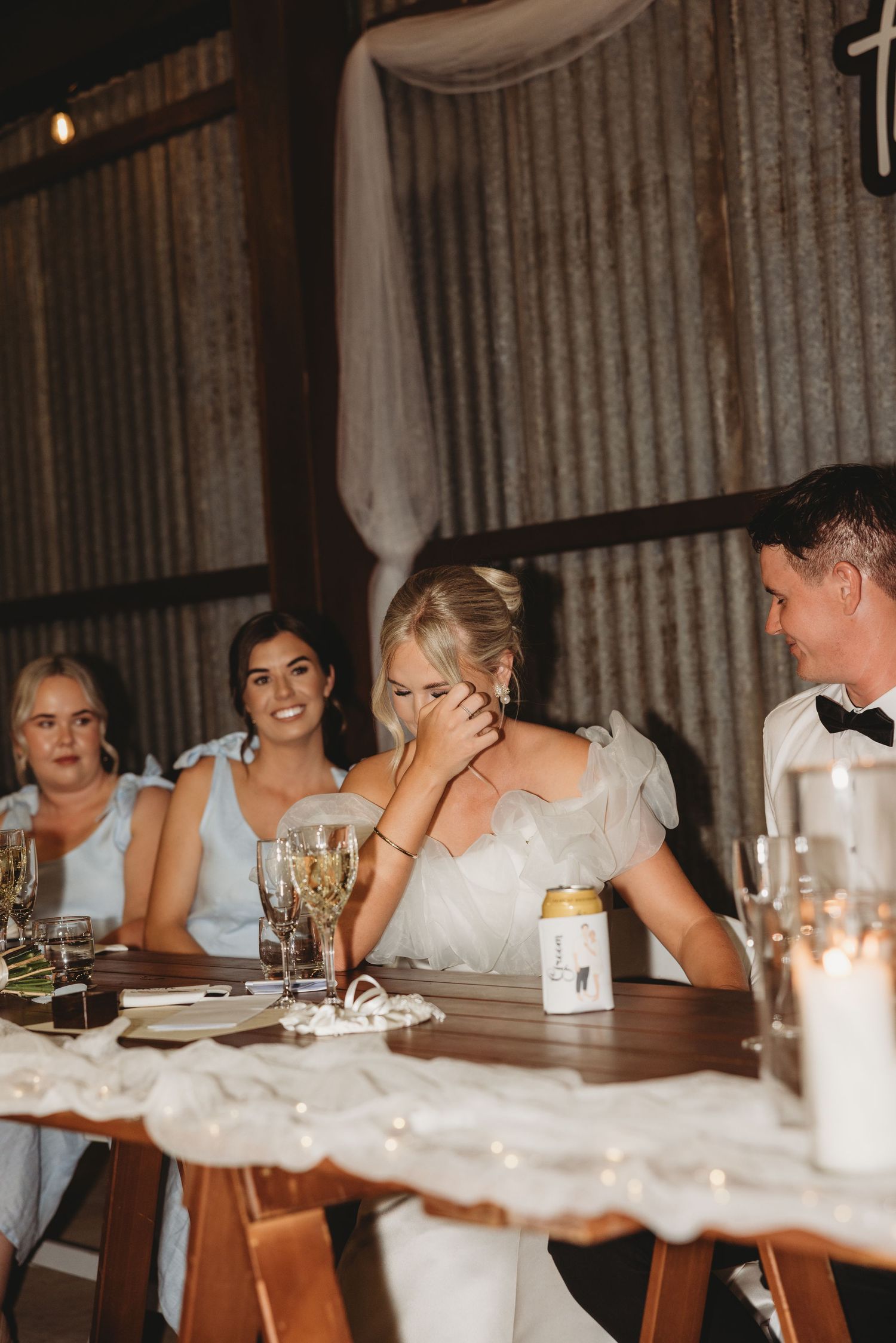 Wedding reception dinner scene with guests seated at a rustic wooden table with white linens and candles.