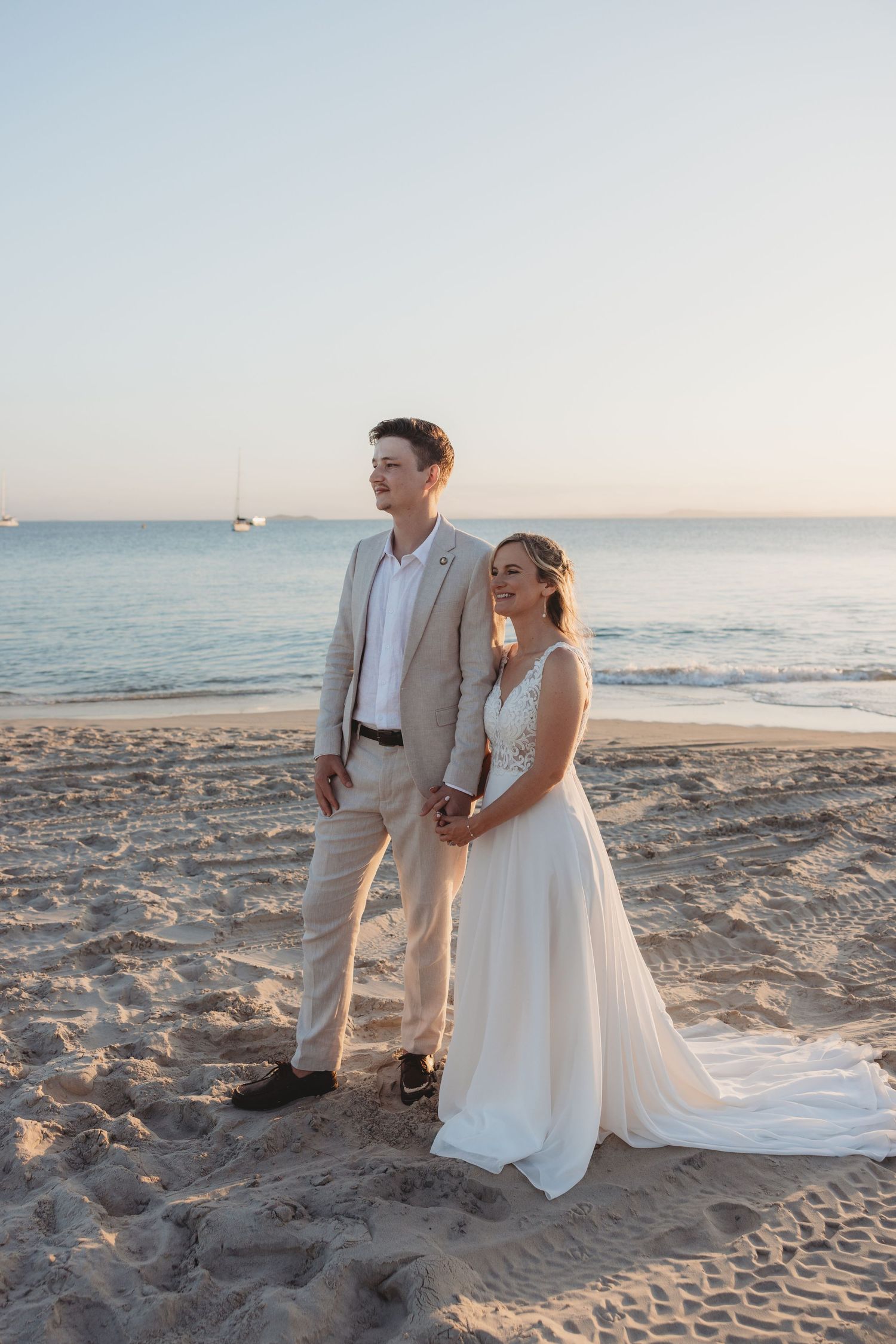 Beach wedding couple poses on sandy shore with ocean waves in background at sunset.