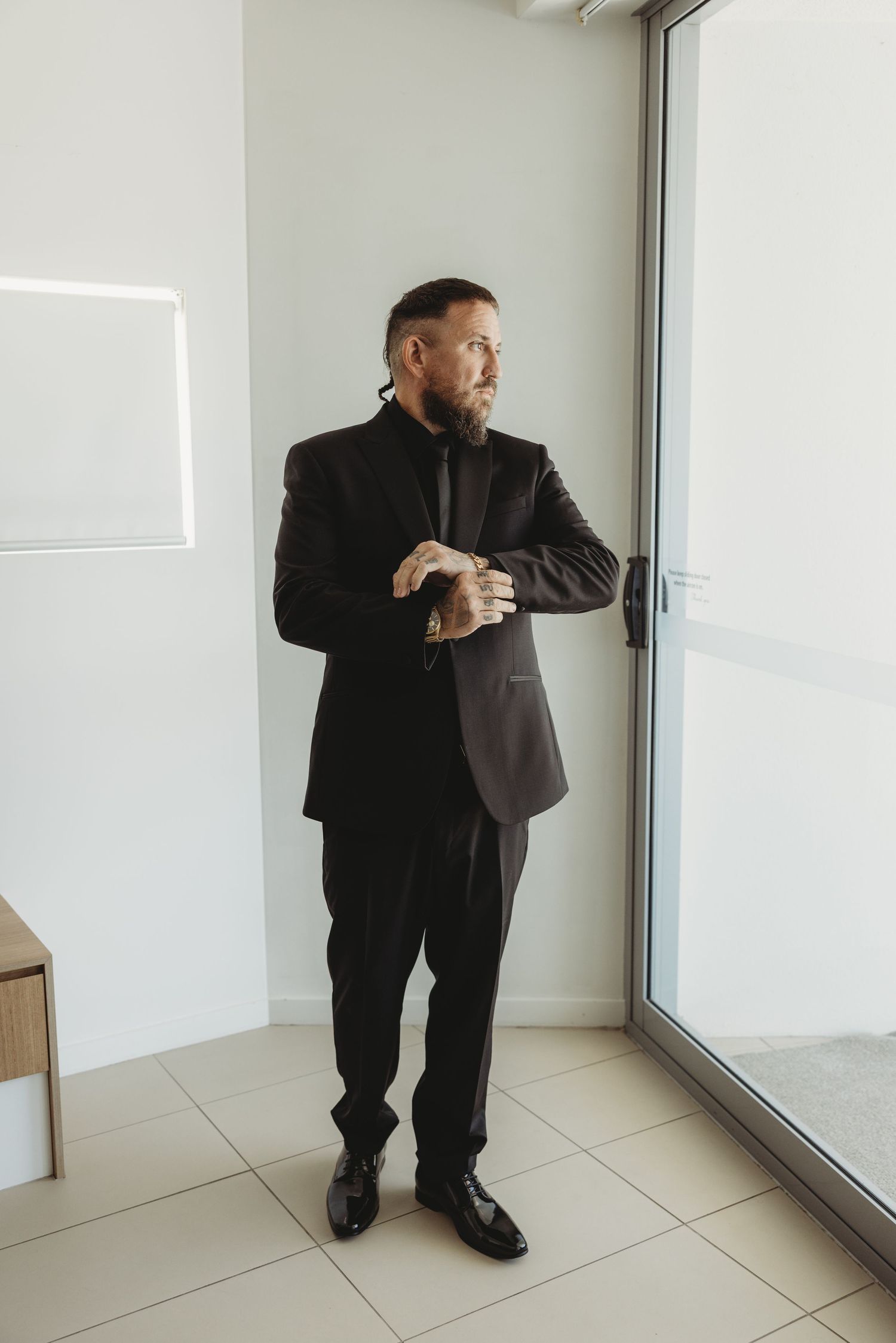 Person in black suit peering through window in minimalist white interior setting.