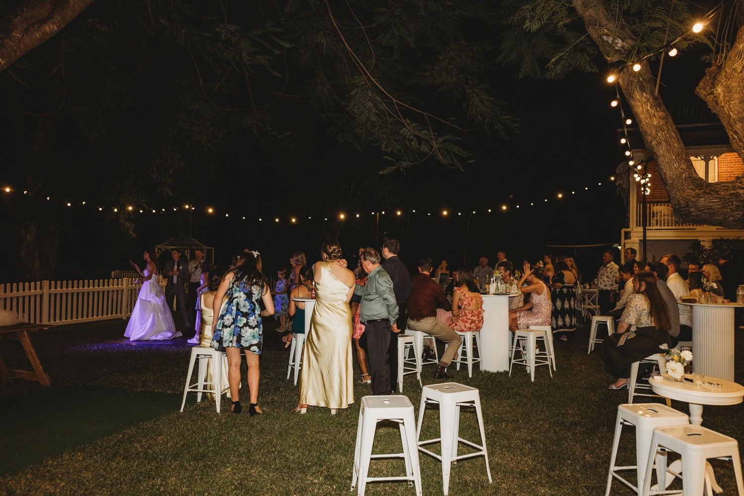 Outdoor wedding reception setup with string lights and white chairs under trees.