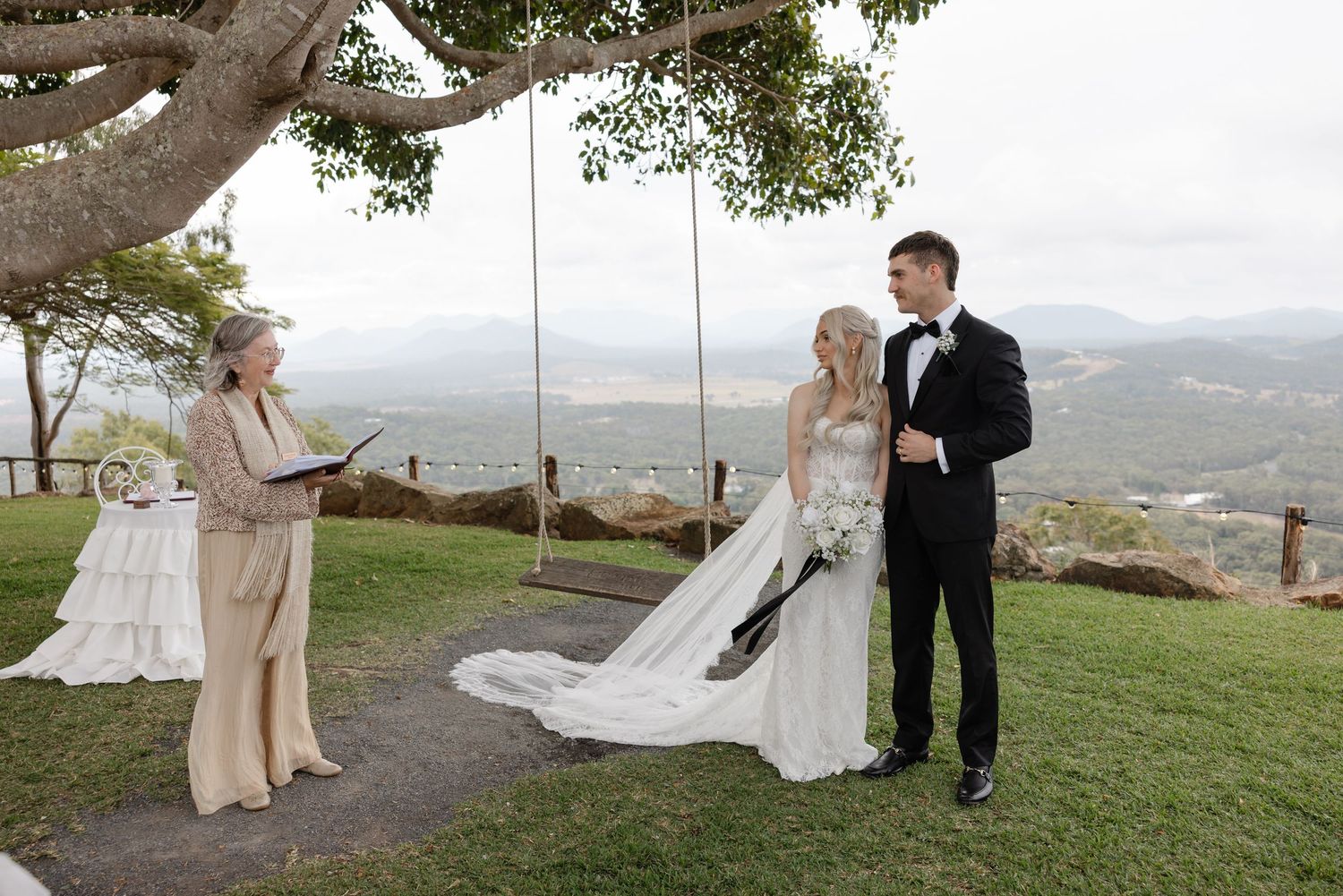 Bride and groom exchange vows outdoors with officiant, scenic valley views and rustic tree swing backdrop.
