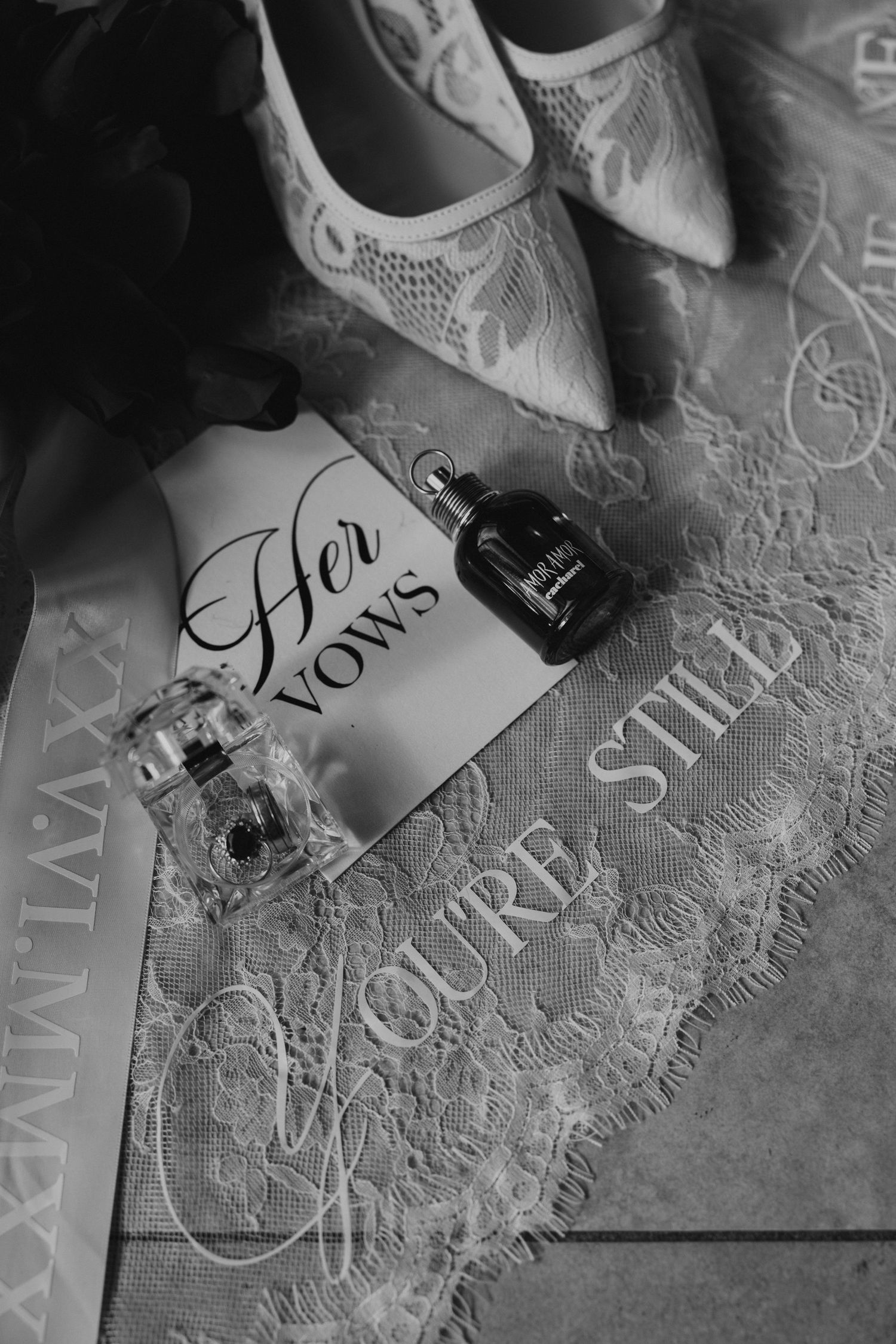 Black and white flat lay of bridal vows card, perfume bottles, lace garter, and white heels for a wedding day.