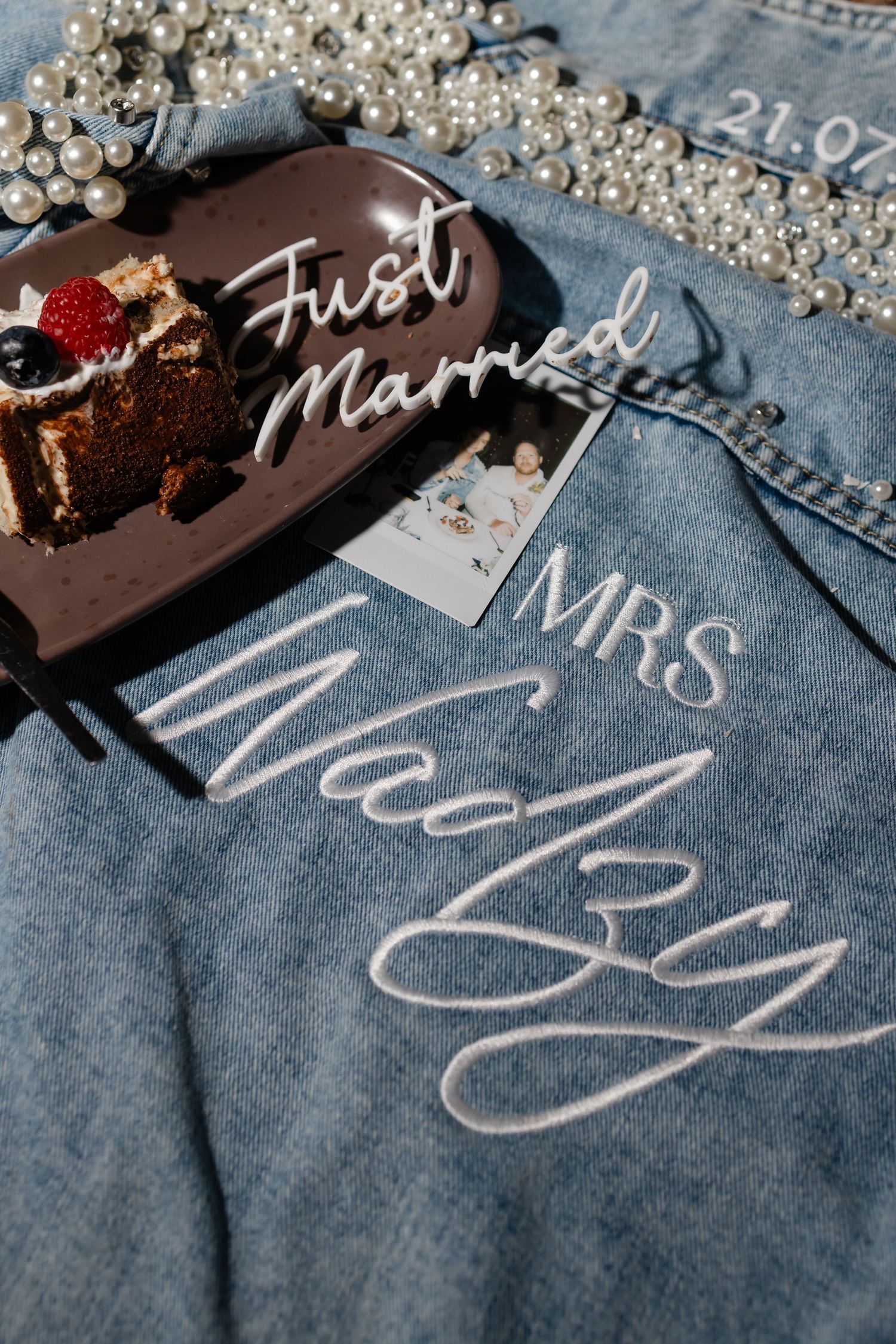 Custom denim jacket embroidered with MRS and last name, pearl necklaces, heart plate with cake slice, and Just Married acrylic sign.