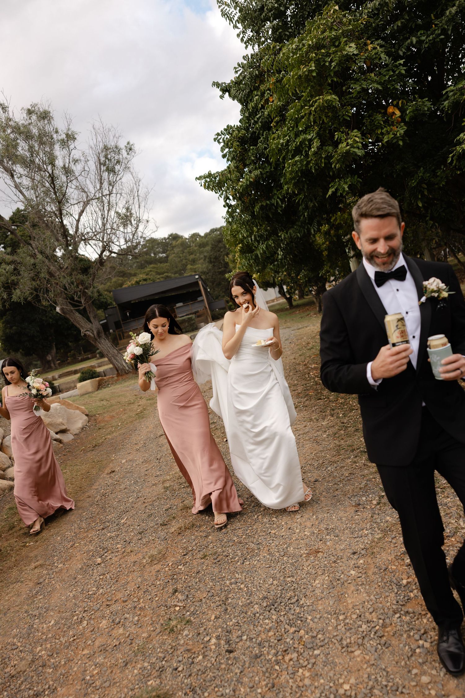 Groom in black tuxedo leads bride in white gown and bridesmaids in pink dresses along a gravel path outdoors.