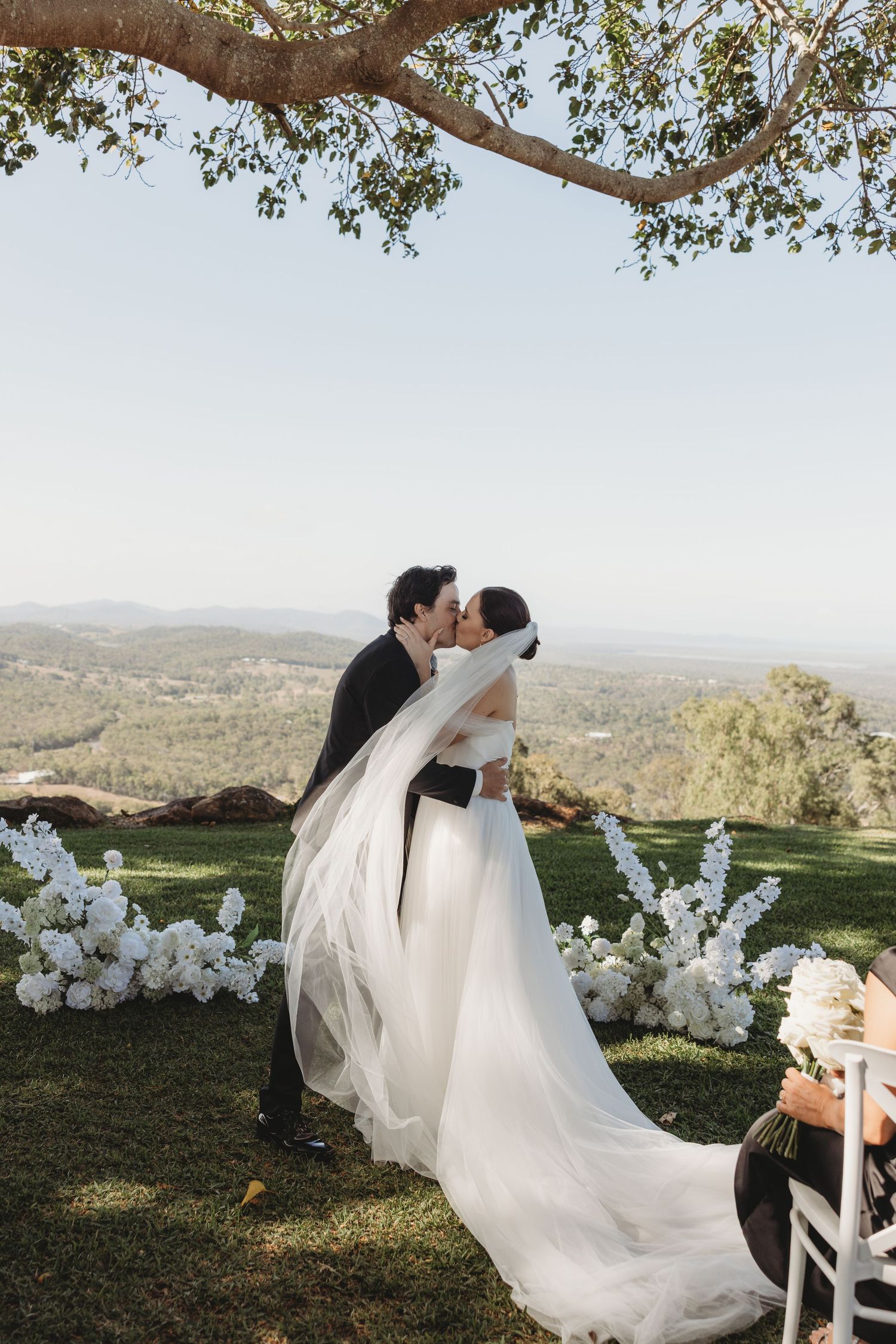 Wedding couple shares romantic moments under a tree branch with white floral decorations and mountain views.