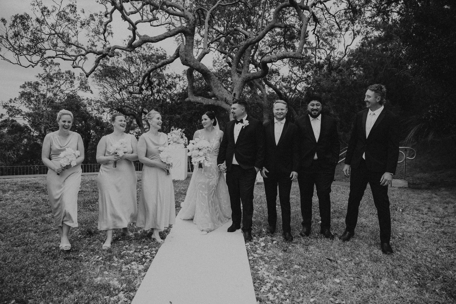 Black and white photo of a wedding party lined up on a white aisle runner outdoors.