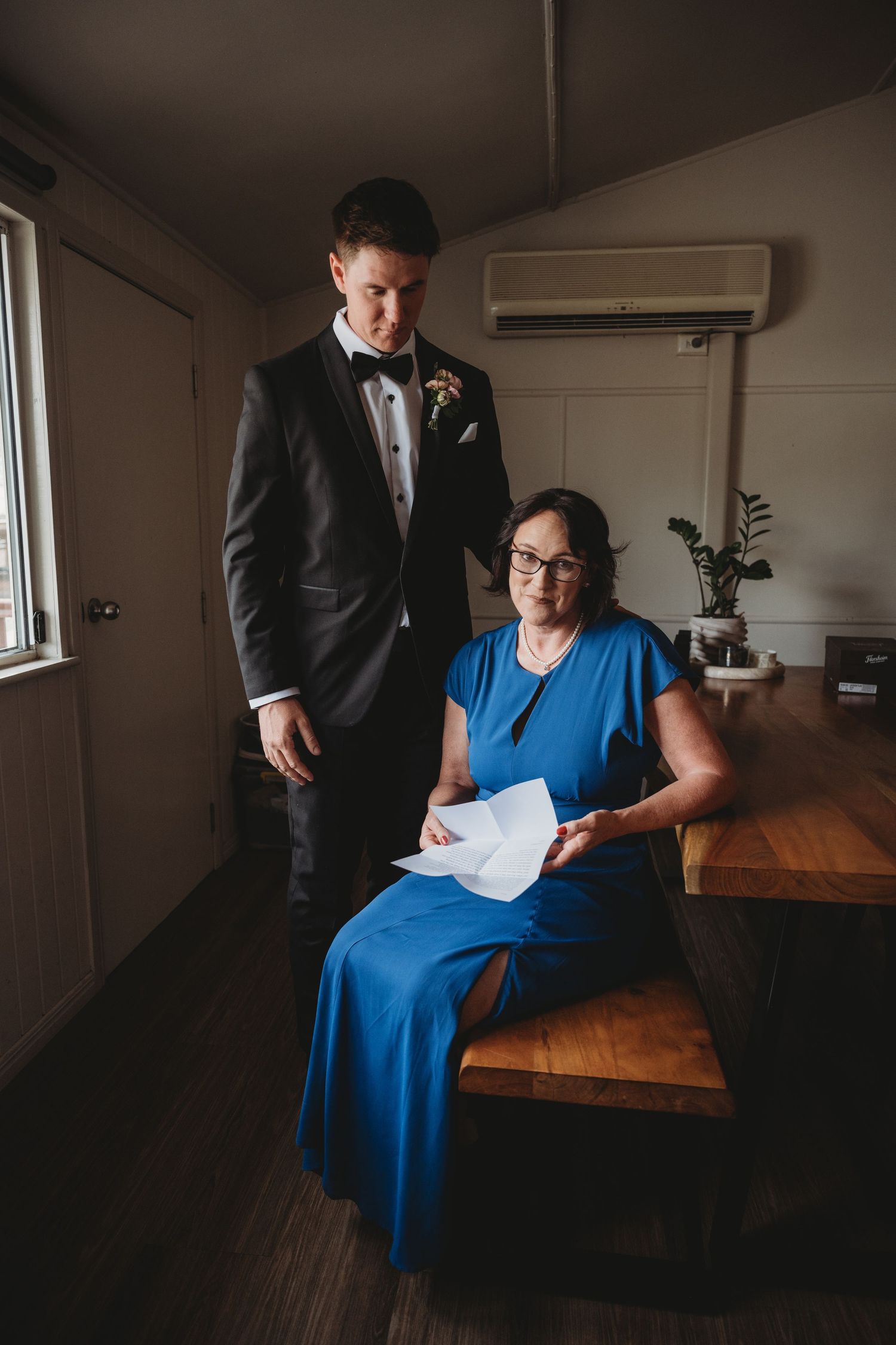 Wedding party member in blue dress sits thoughtfully by window light.