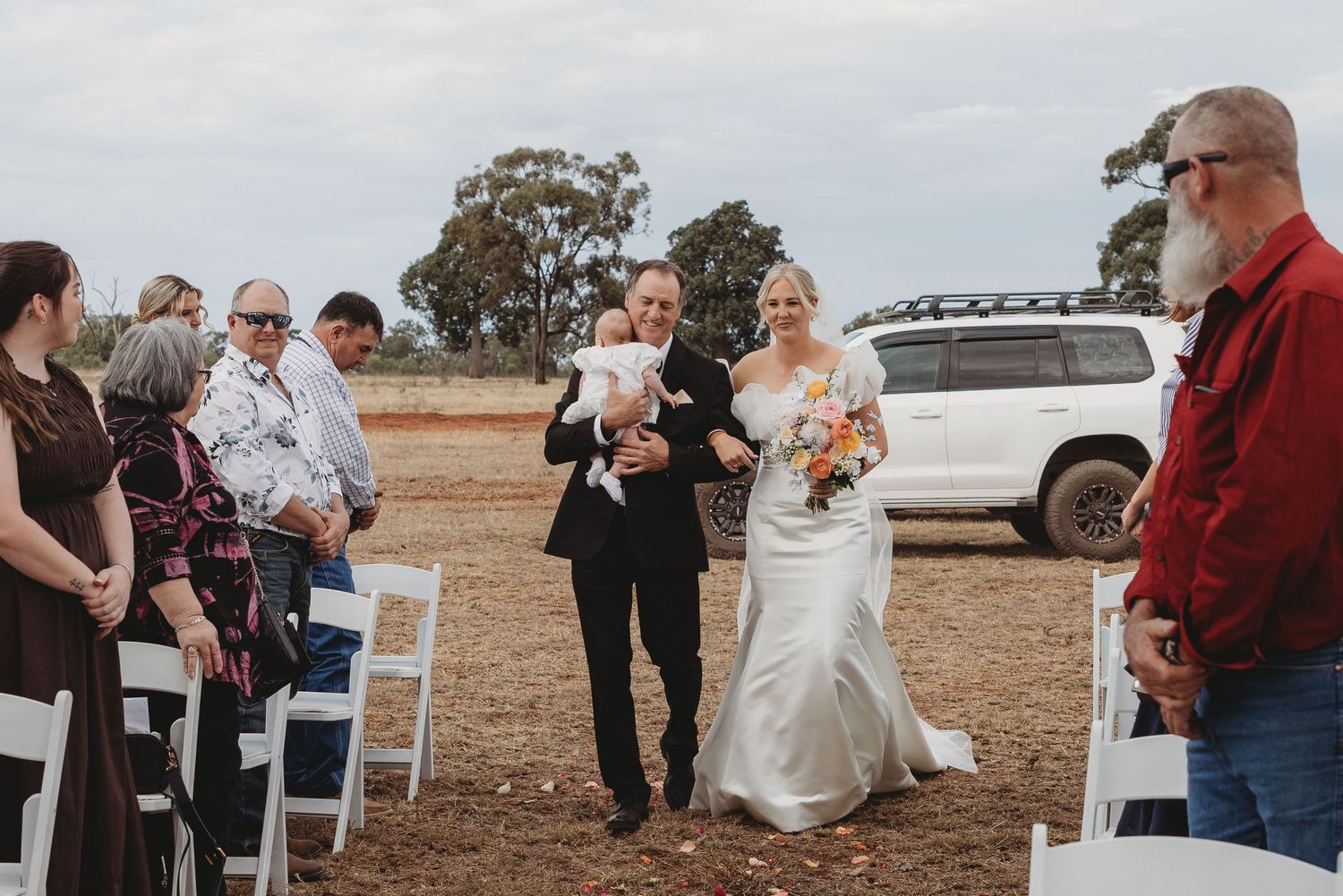 Wedding guests gathered outdoors under a cloudy sky for a countryside celebration.