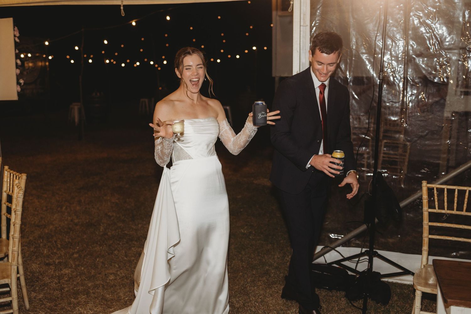 Bride and groom make grand entrance holding drinks at outdoor evening wedding reception.
