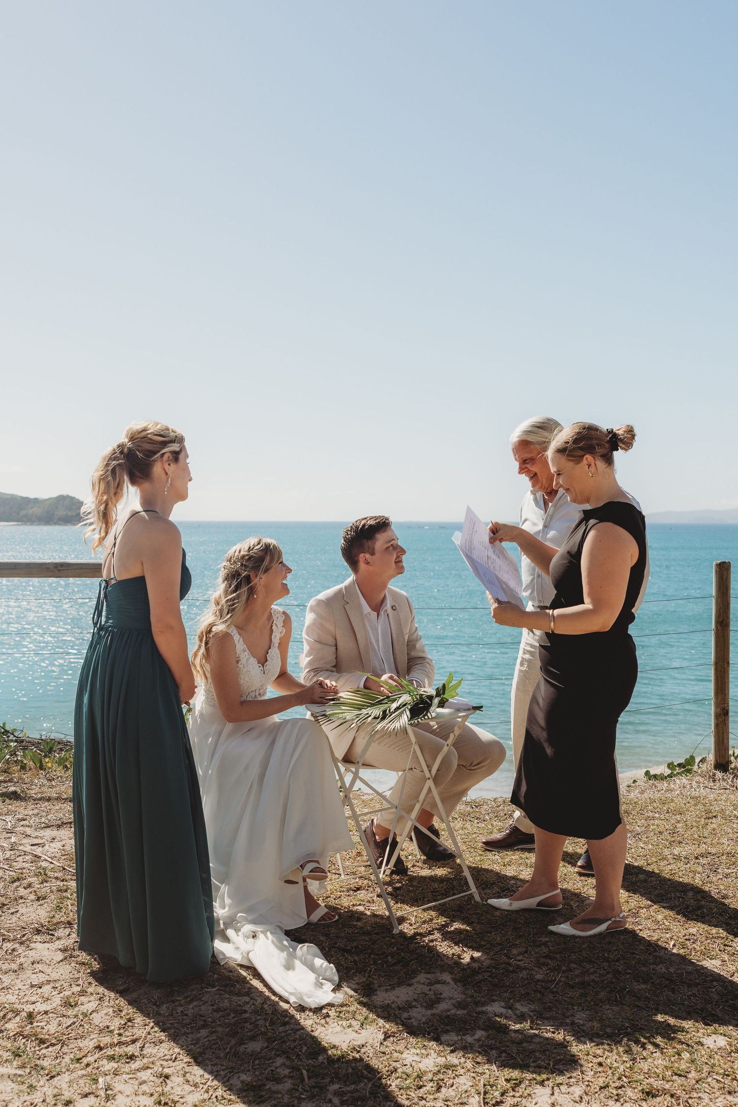 Wedding guests and couple gather on coastal cliff overlooking bright blue ocean.