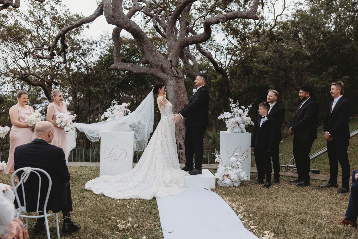 Wedding ceremony taking place in a natural setting with guests seated beneath a sprawling tree.