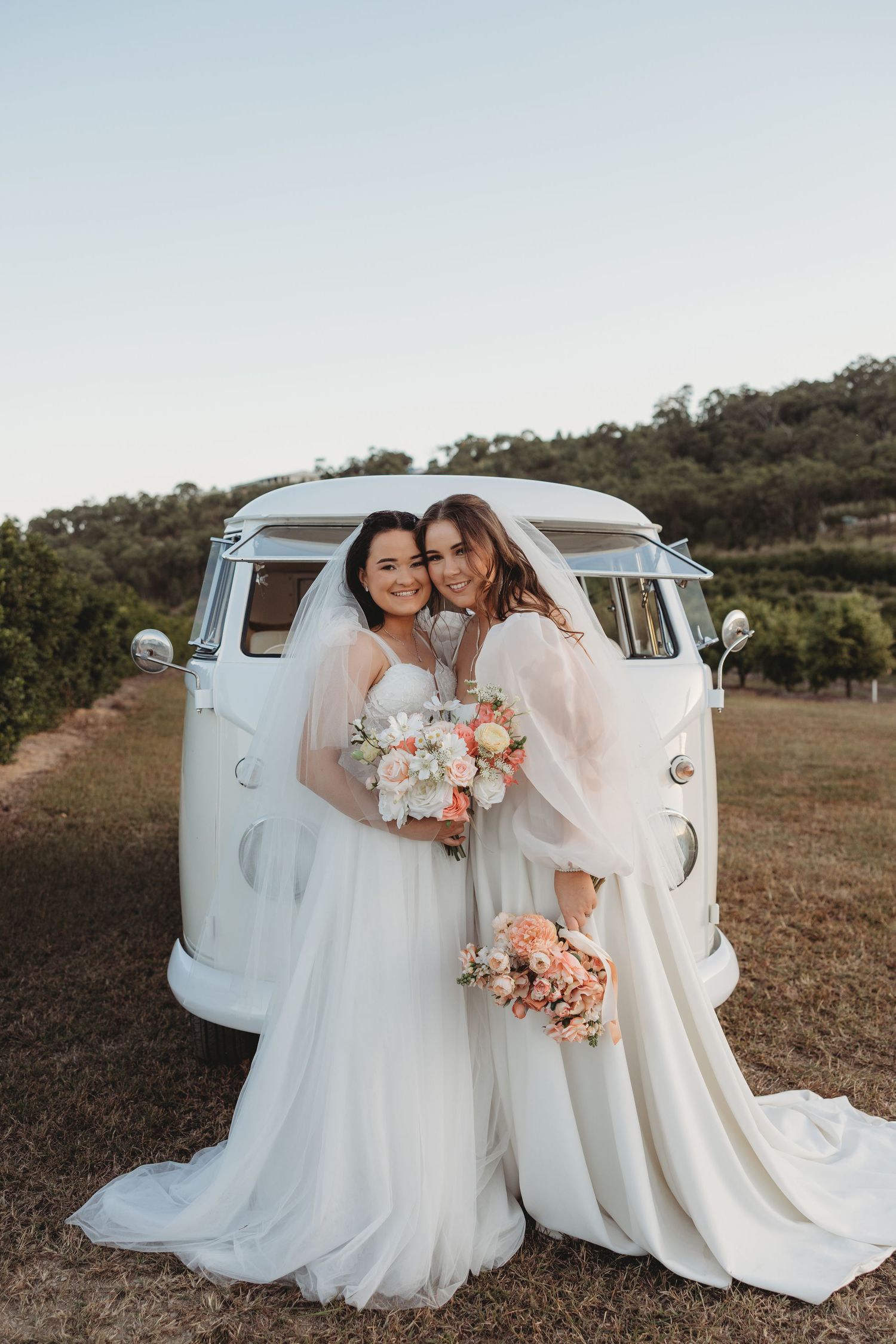 Two people in wedding dresses posing in front of a classic white Volkswagen van.