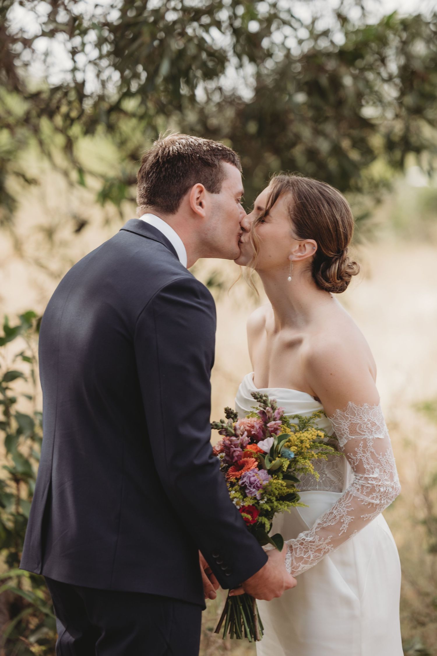 A bride and groom share a kiss outdoors, bride holding a vibrant wildflower bouquet in lace wedding dress.