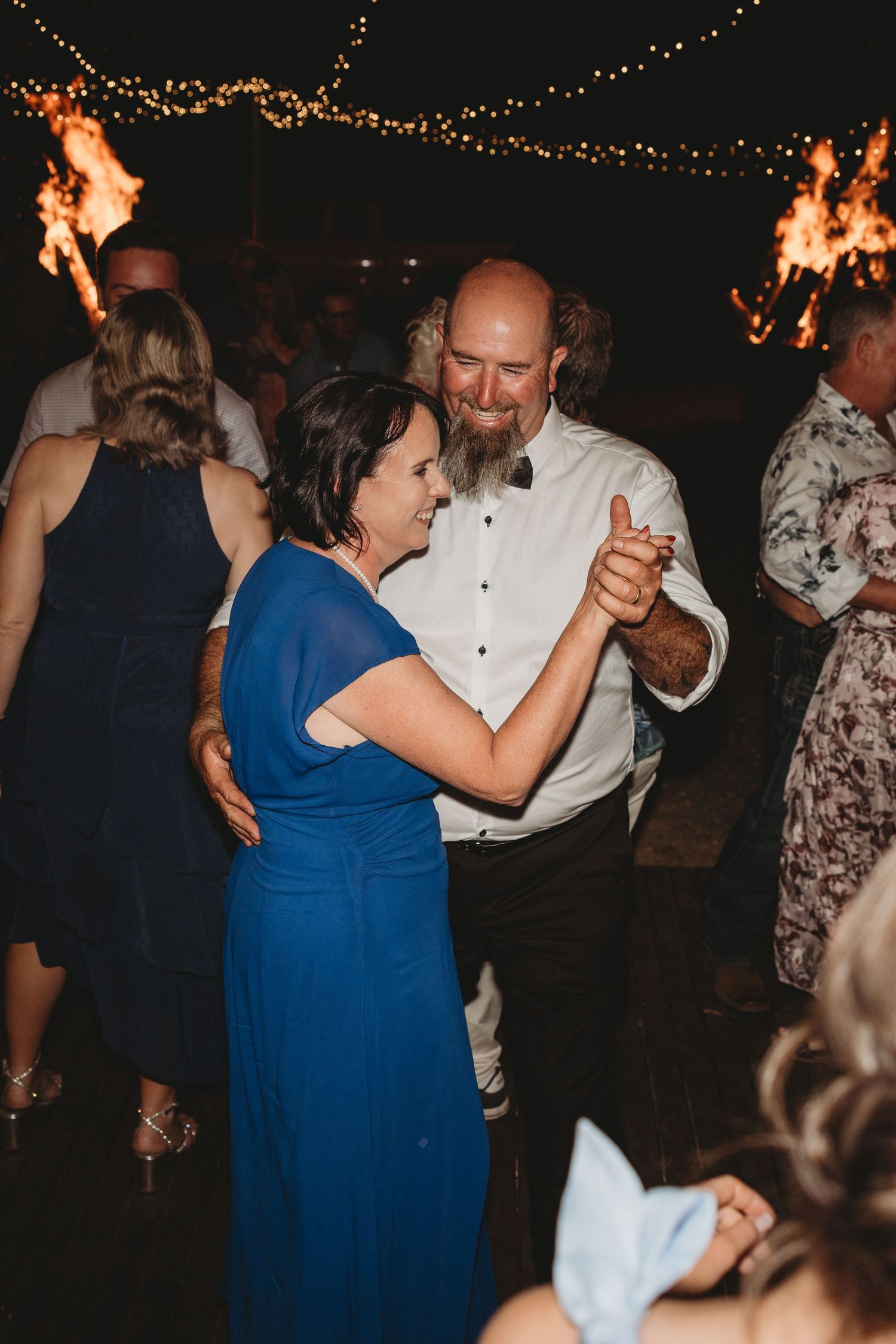 Guests dancing at wedding reception with string lights and fire torches in background.