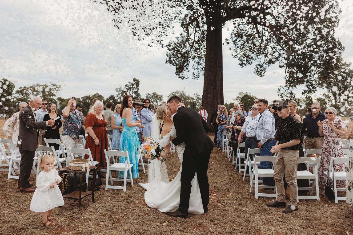 Wedding guests seated in white chairs applaud during outdoor ceremony under trees.