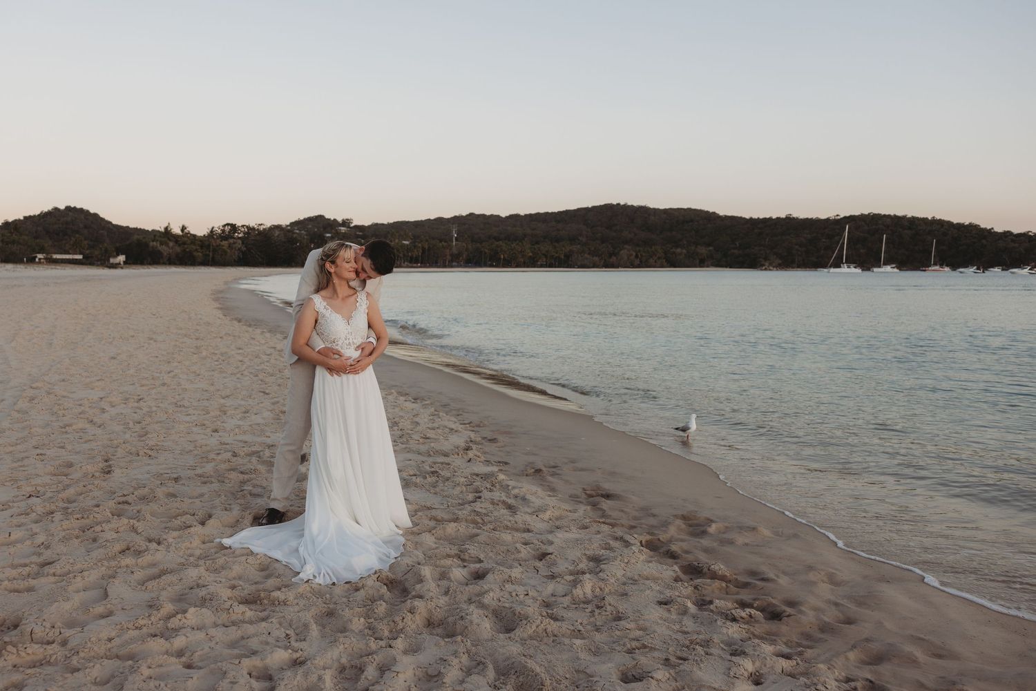 A couple shares a romantic moment on an empty beach with sailboats visible in the distance.