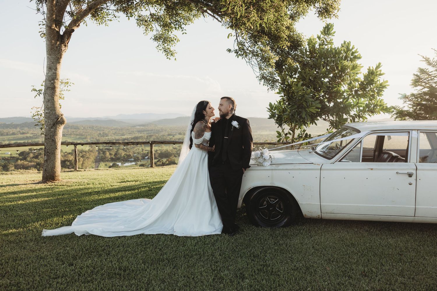 Wedding couple posing near a classic white car on a scenic hilltop during sunset.