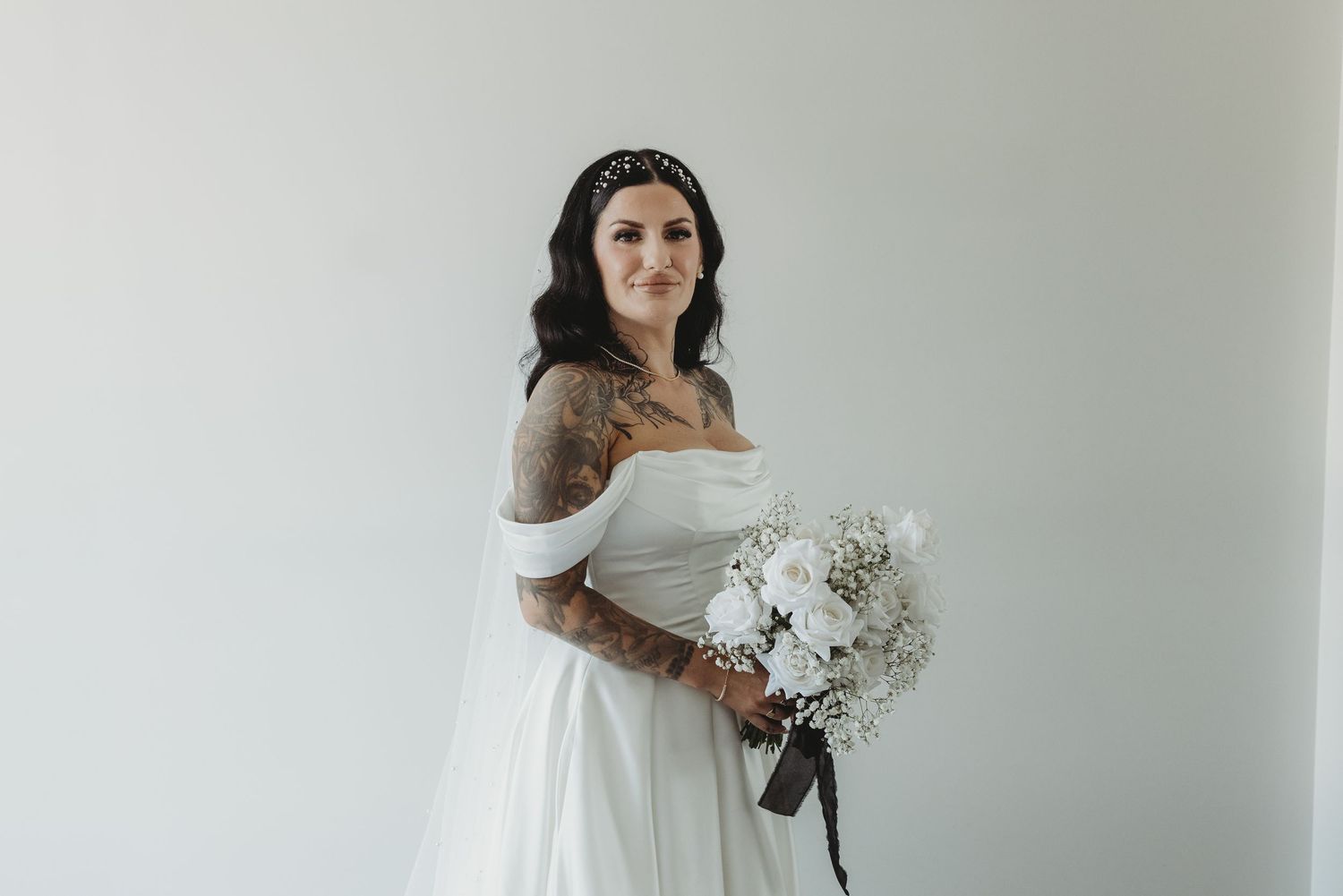 A bride in a white off-shoulder wedding dress poses with a delicate bouquet of baby's breath flowers.