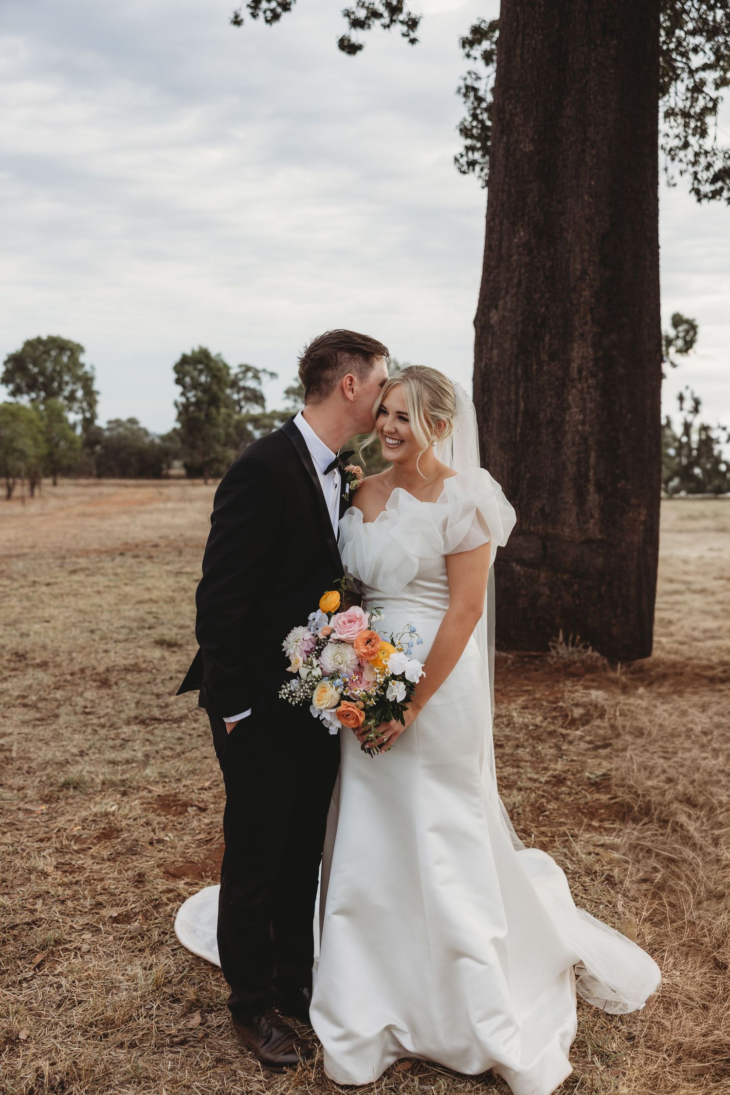 Romantic outdoor wedding photo with couple holding bouquet beneath towering pine trees.