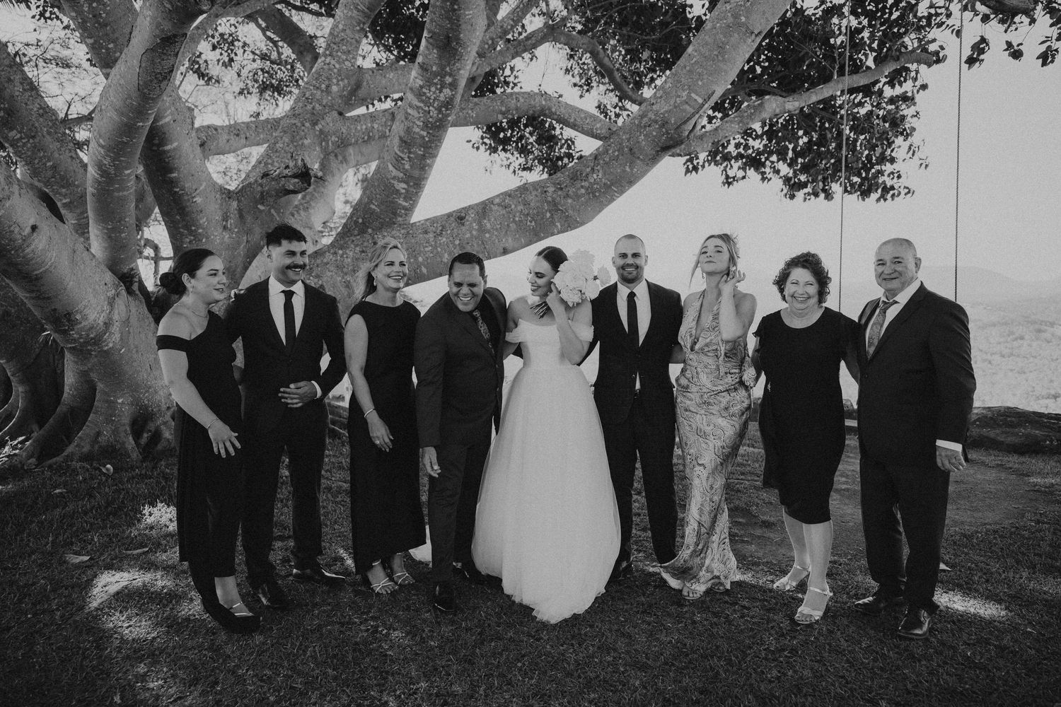 Large wedding party gathers for group photos under a majestic tree.