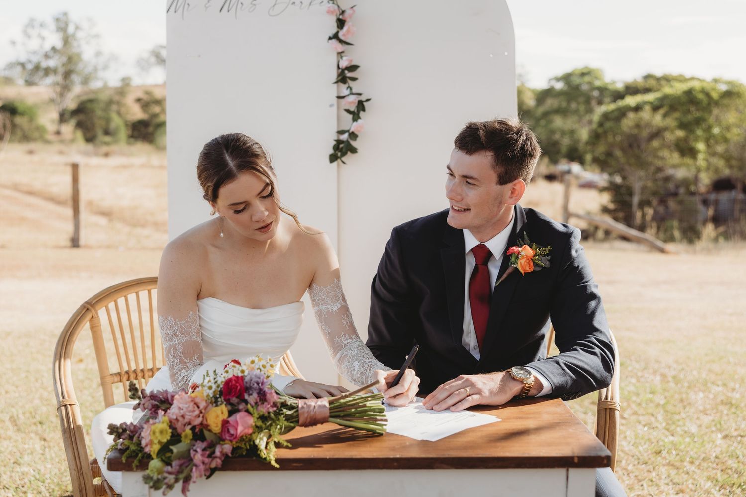 Bride and groom signing wedding register at outdoor ceremony table with colorful bouquet.
