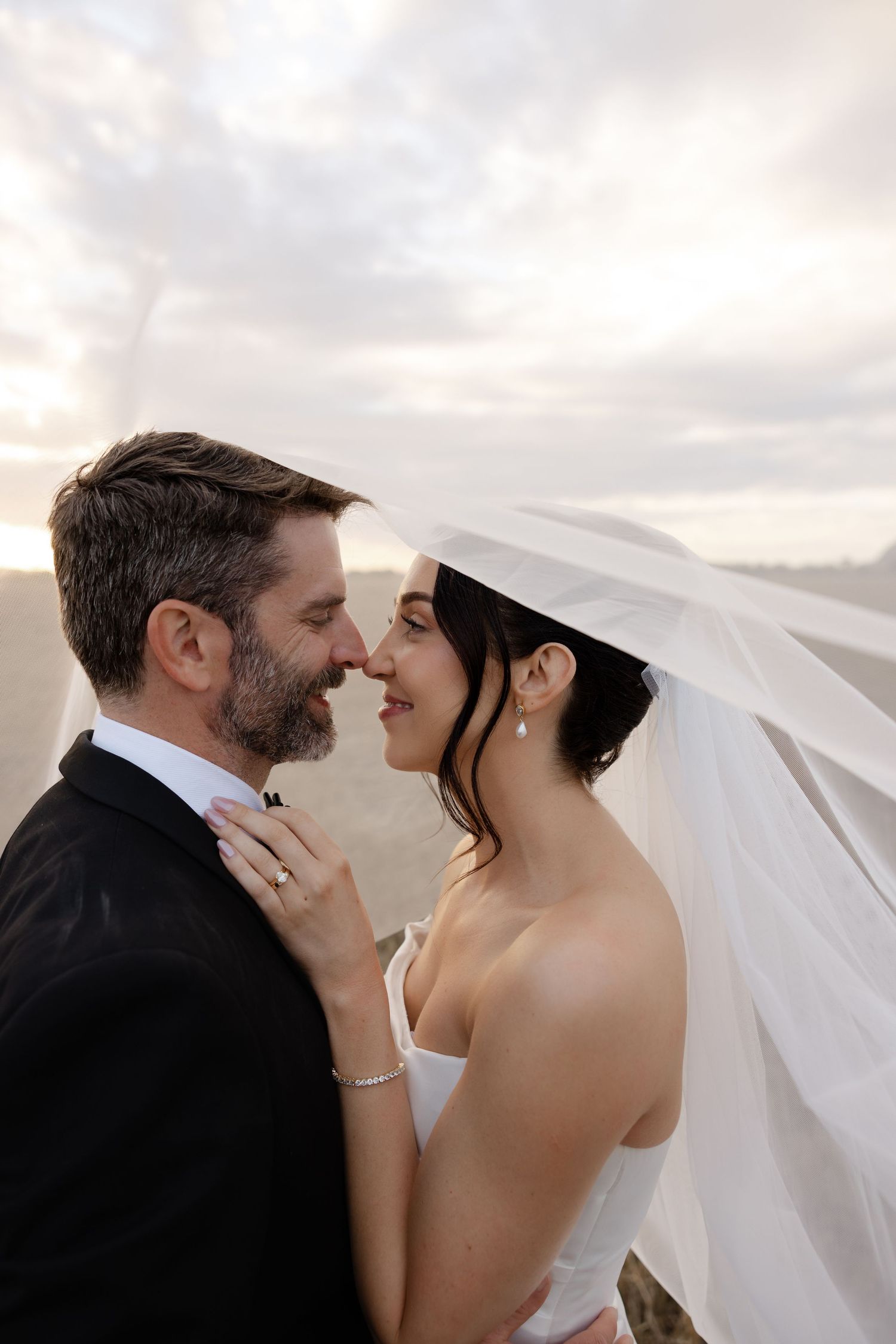 A bride and groom share a romantic moment under a flowing veil at sunset, smiling at each other outdoors.
