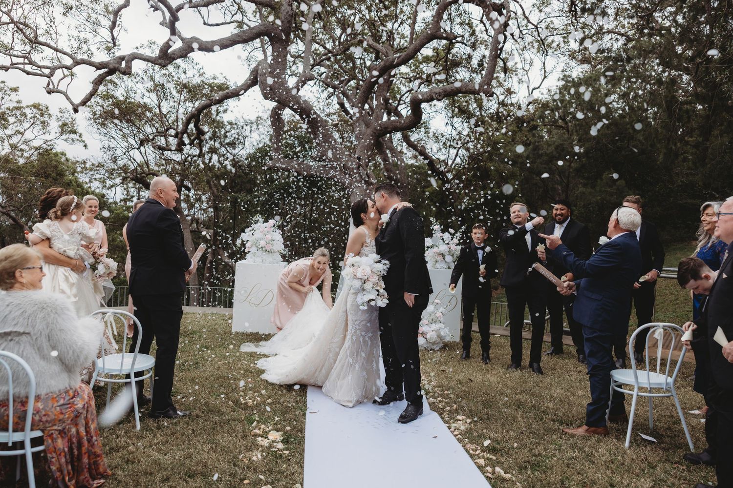 Outdoor wedding ceremony taking place under trees with guests seated on white chairs.