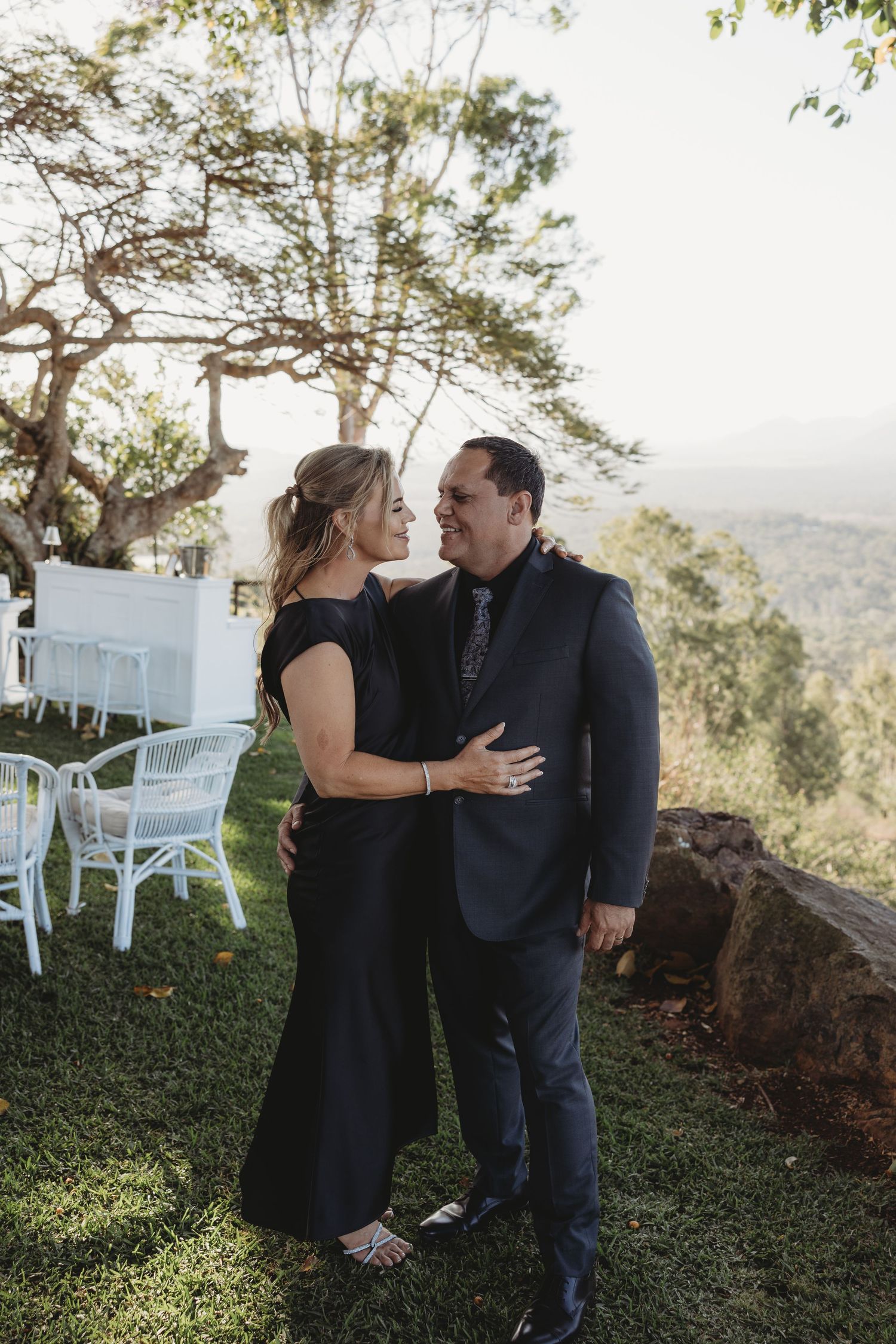 A couple sharing romantic moments on a grassy hillside with white chairs in the background.