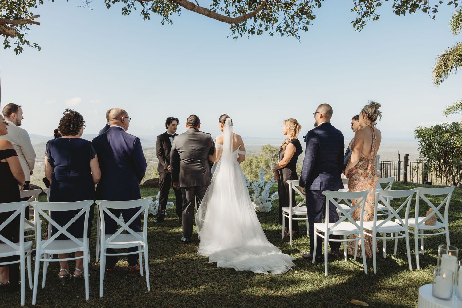 Wedding guests seated in white chairs on a grassy hillside overlooking a scenic valley landscape.