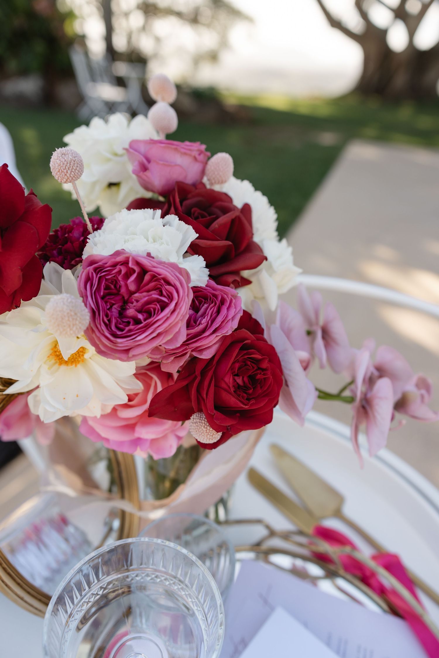 Elegant wedding table centerpiece with red, pink, and white roses in a glass vase with gold cutlery.