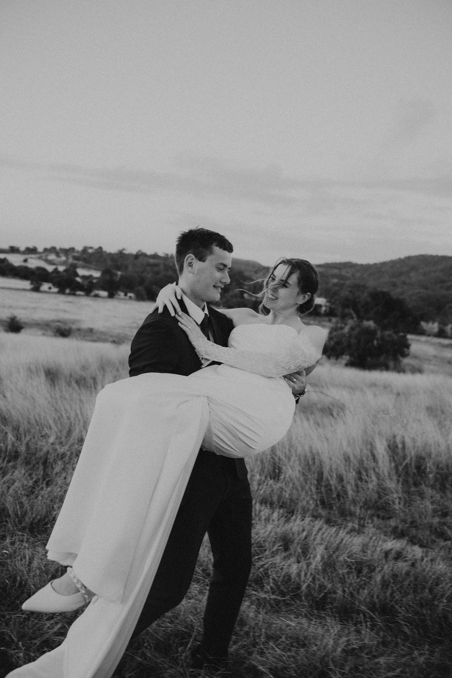 Groom carries laughing bride through golden field in classic black and white wedding photo.