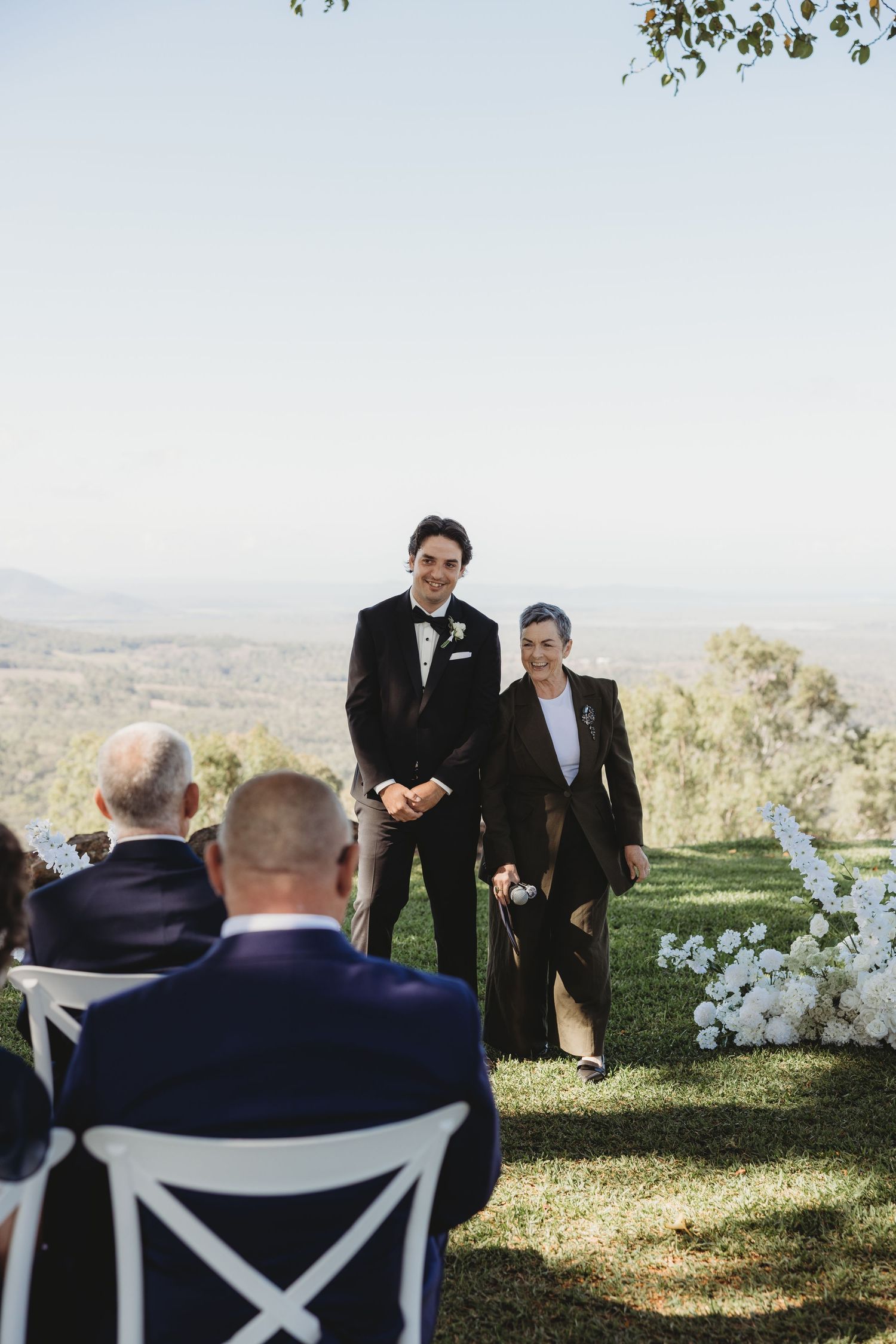 Outdoor wedding ceremony setup with men in suits standing near white chairs on a scenic hilltop.