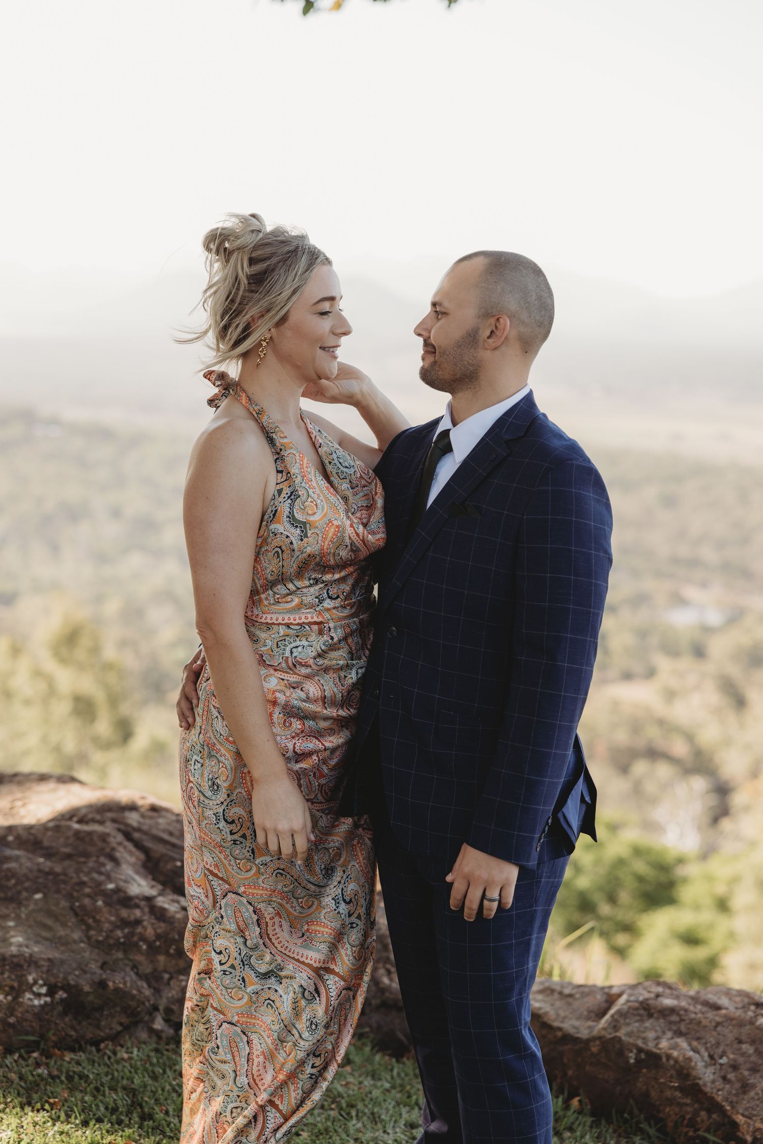 A couple in formal attire share an intimate moment on a scenic overlook with mountains in the background.