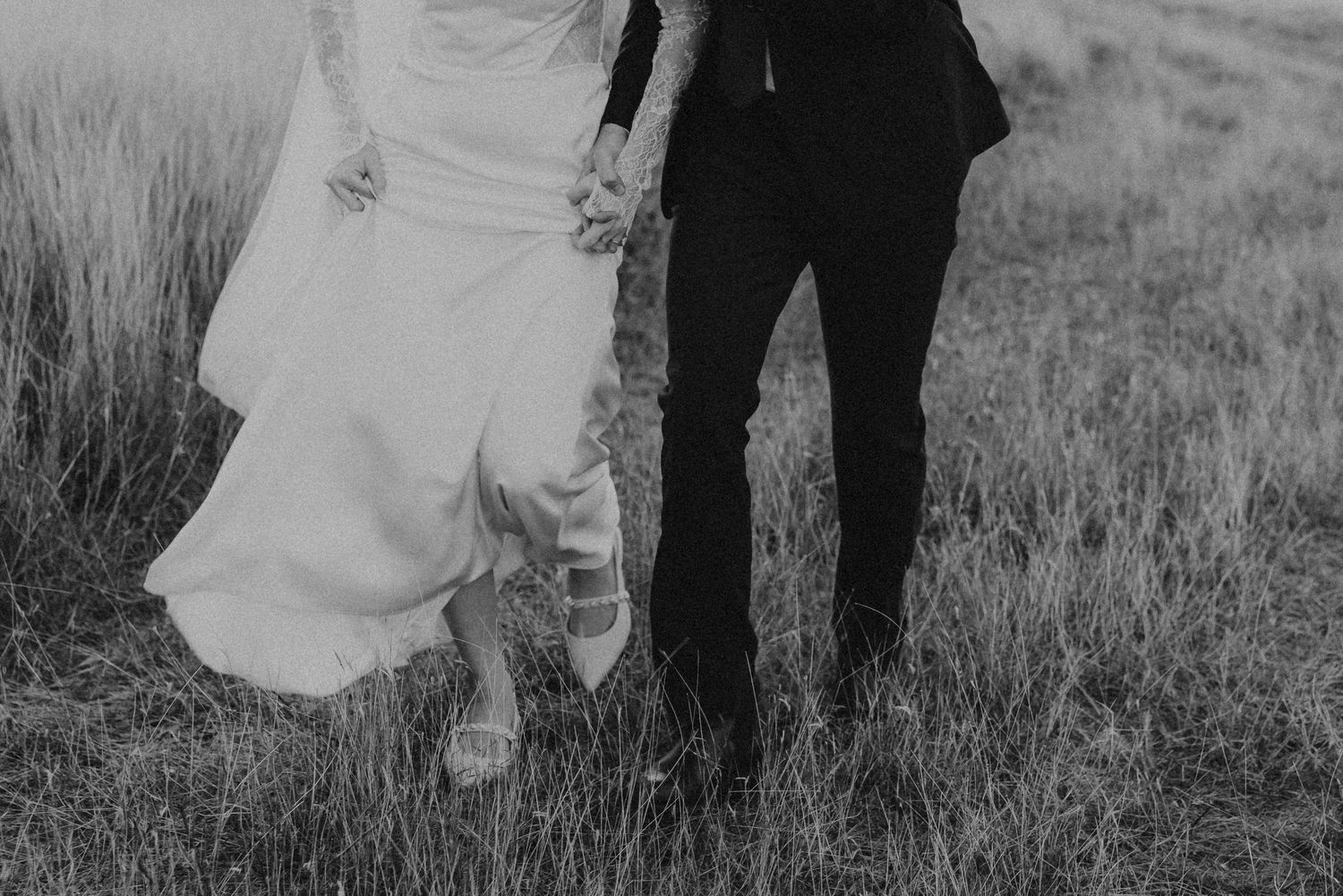 Black and white photo of bride and groom holding hands walking through tall grass at their wedding.