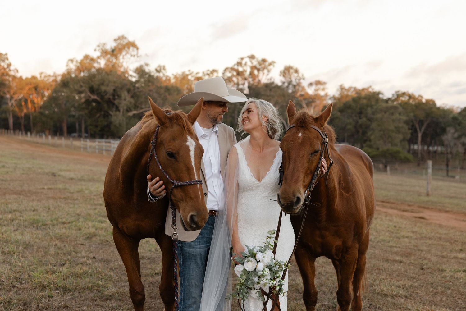 Bride and groom stand between two chestnut horses at golden hour on a rustic farm, bride holding white bouquet.