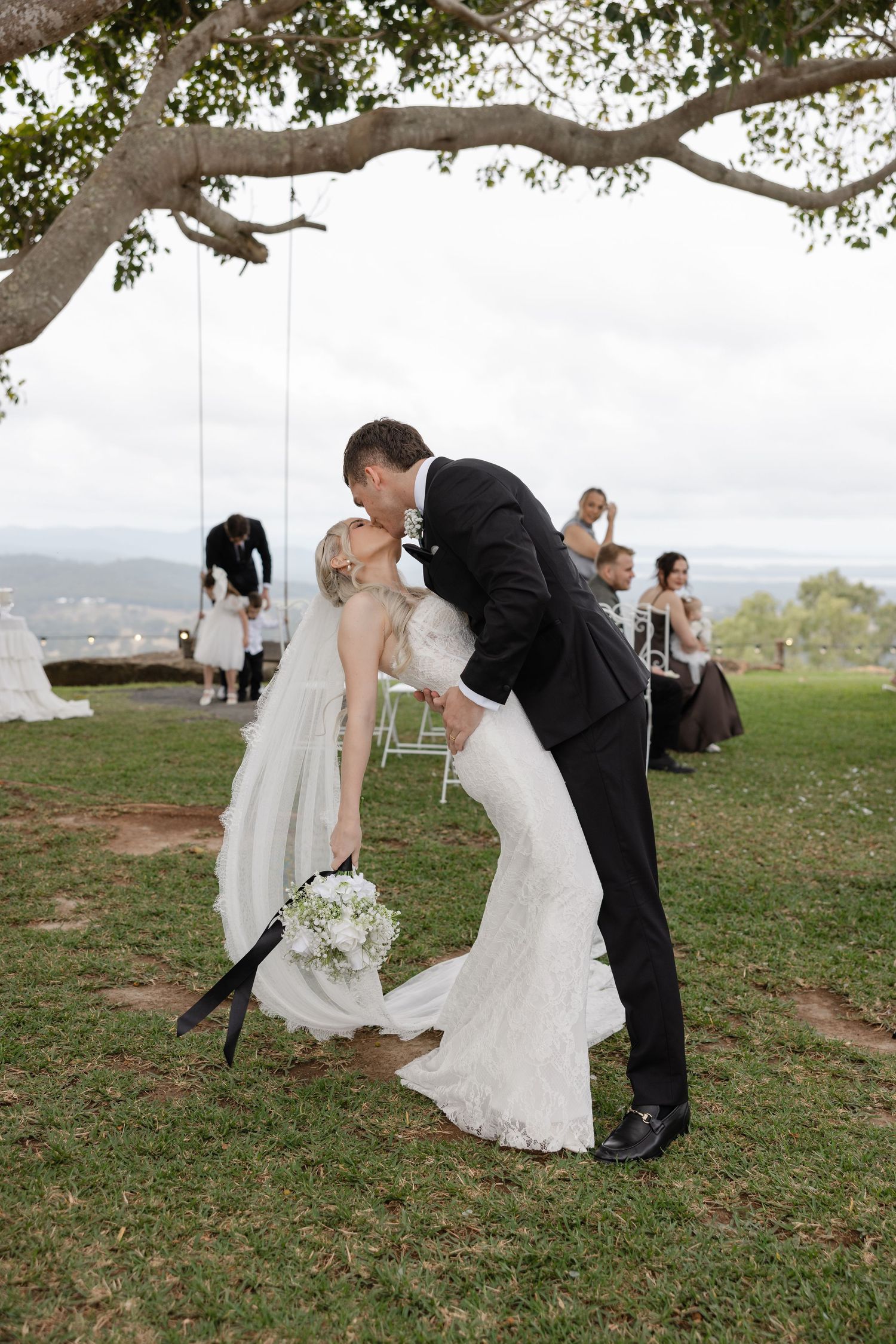 Bride and groom share a romantic dip kiss on green lawn during outdoor wedding ceremony with guests in background.