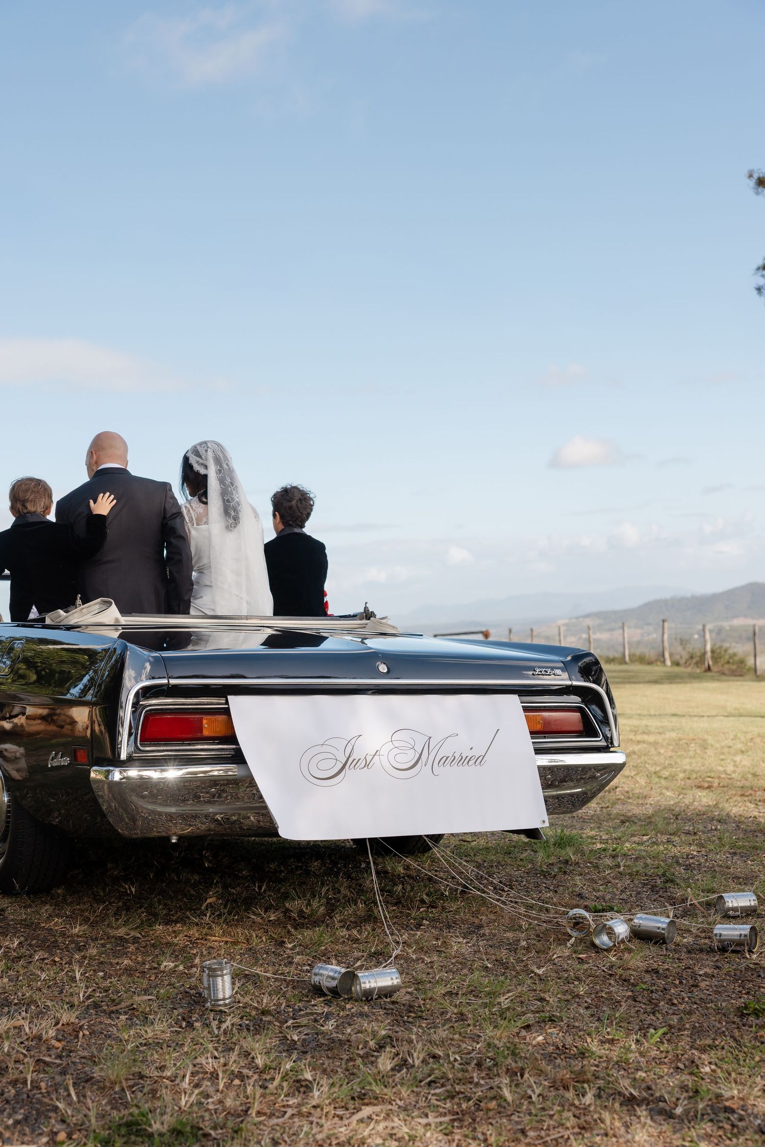 Newlyweds stand by a classic convertible car with a Just Married sign and tin cans tied to the bumper.